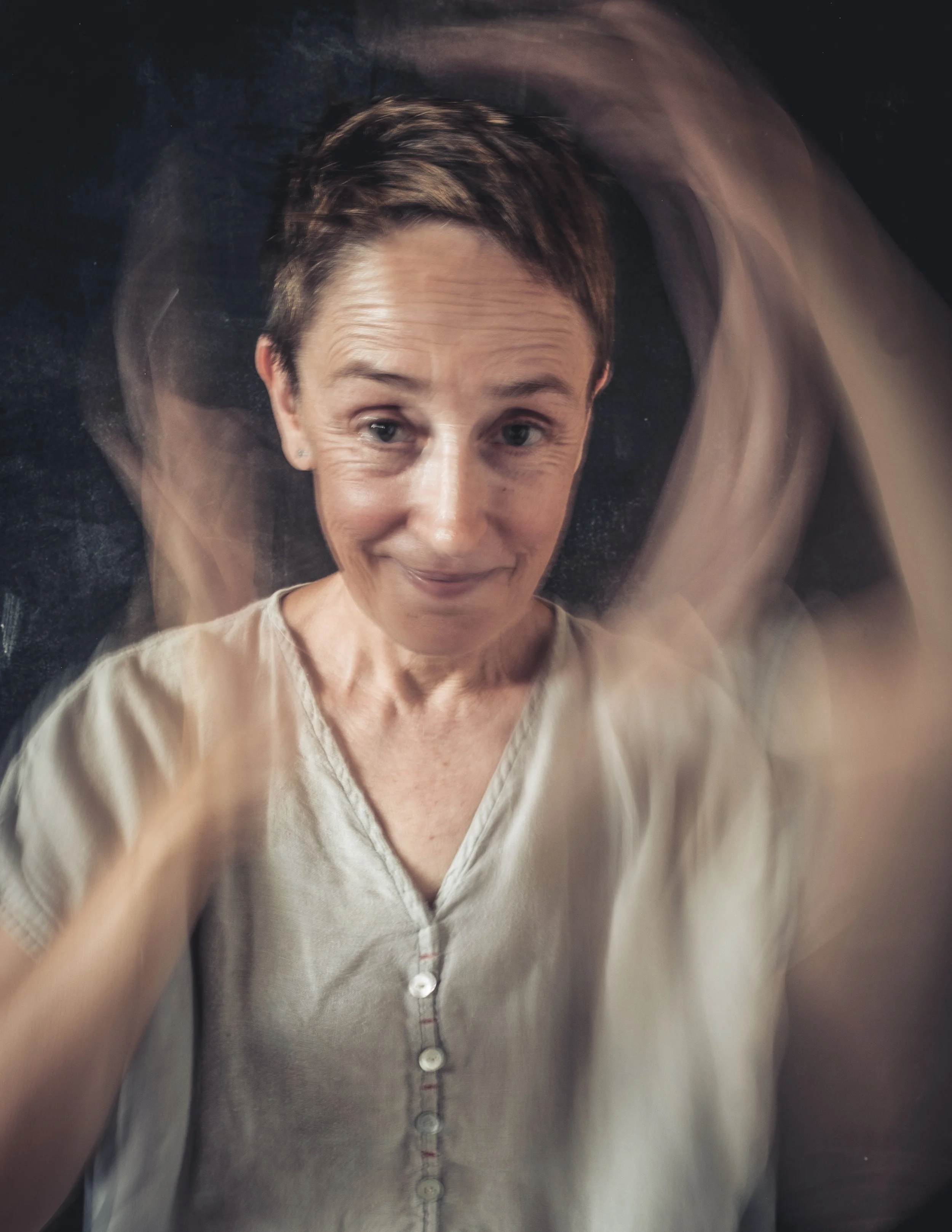 Eine Frau mit kurzen braunen Haaren, die sich die Haare mit den Händen berührt, vor einem dunklen Hintergrund. Das Bild wirkt verschwommen, insbesondere um die Hände, durch eine Bewegungsunschärfe.