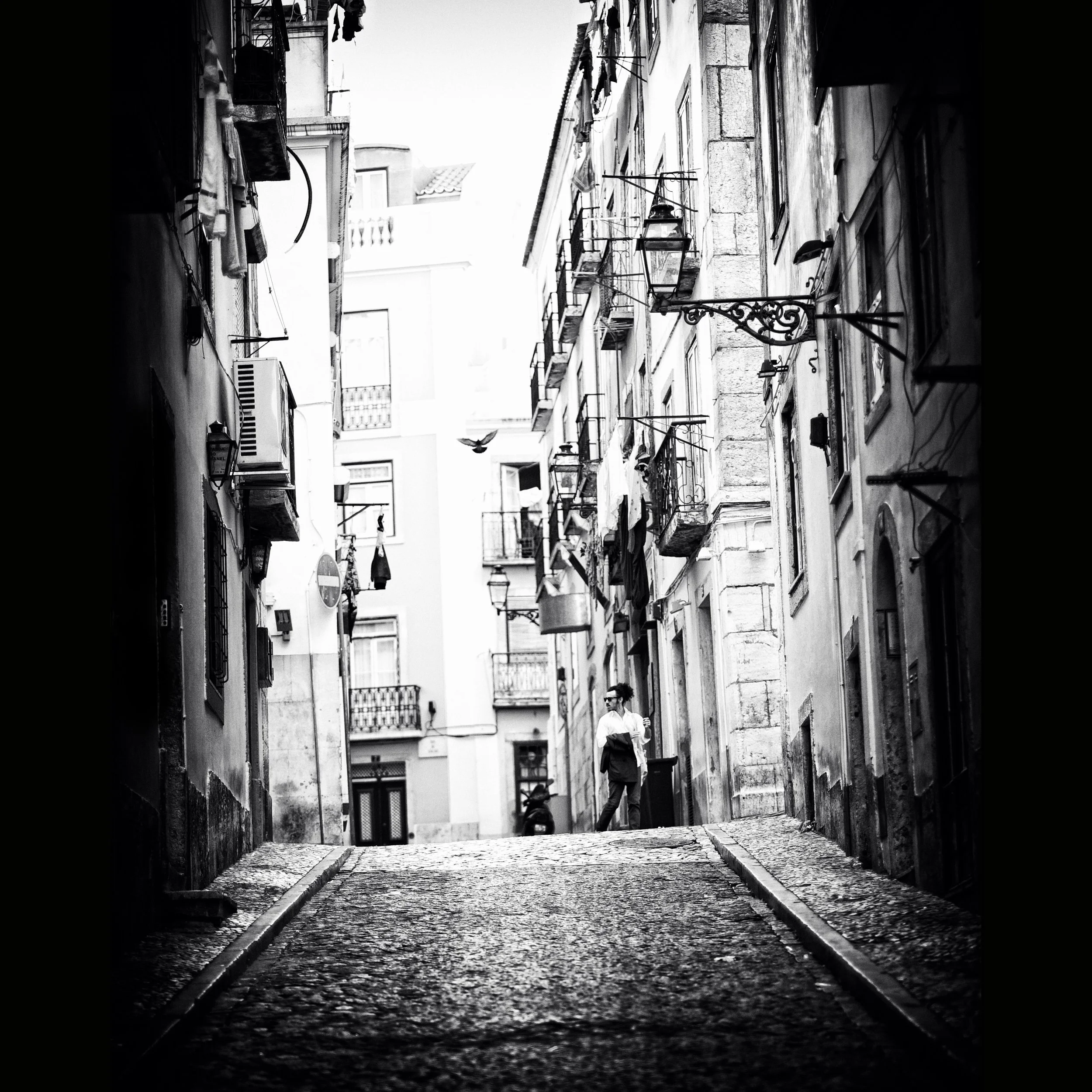 Schmale Gasse mit alten Gebäuden und Balkonen, eine Frau steht in der Mitte und ein Vogel fliegt im Hintergrund, schwarz-weiß