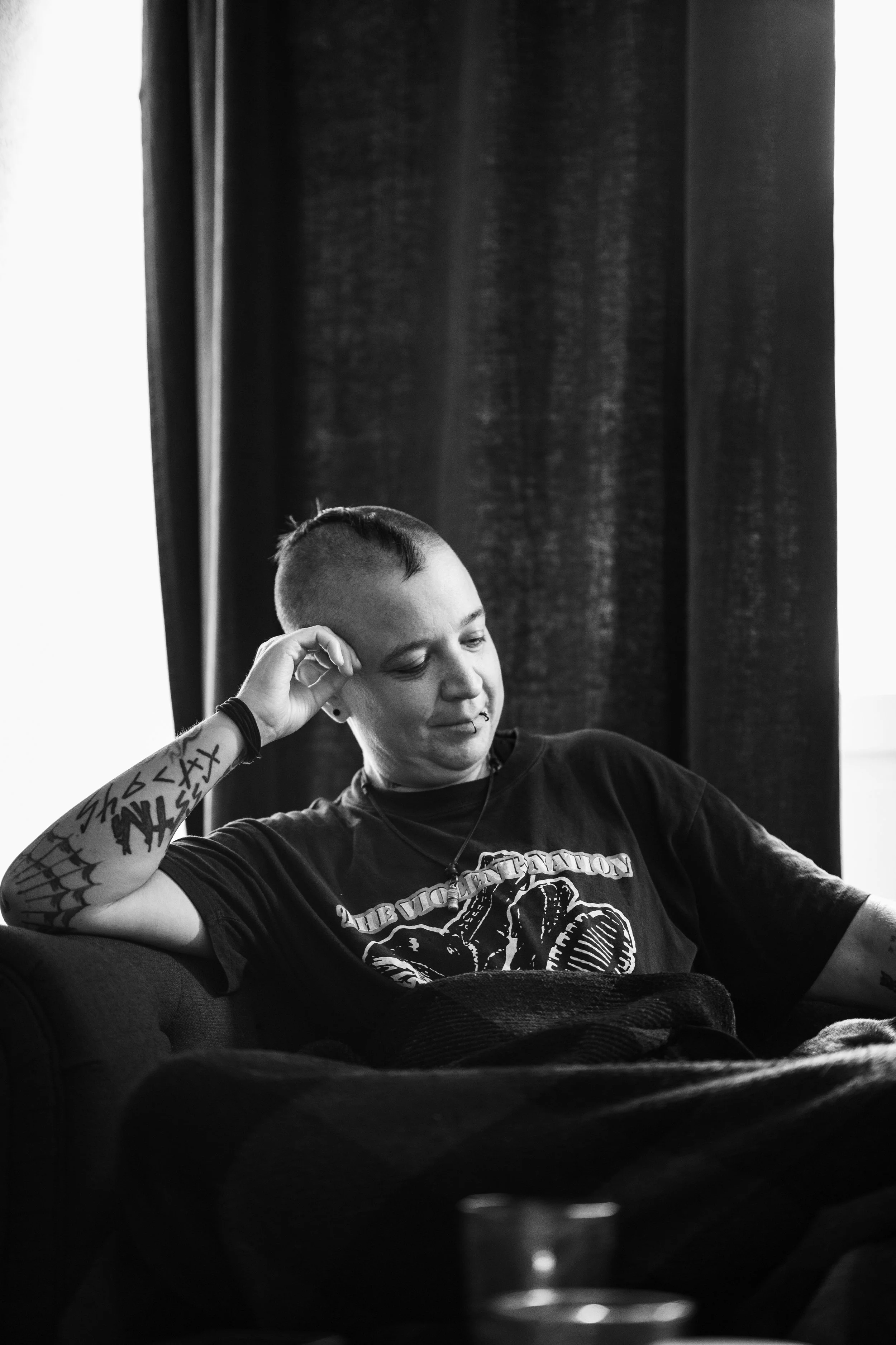 Schwarz-weiße Aufnahme einer Person mit kurzen Haaren, tätowiertem Arm, sitzend auf einem Sofa und entspannt, vor einem Fenster mit dunklen Vorhängen.