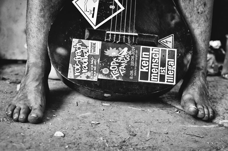 Ein menschliches Bein liegt auf dem Boden, das Bein ist schmutzig und rau. Zwischen den Beinen liegt eine Gitarre mit mehreren Konzert- und Veranstaltungsplakaten auf der Oberfläche.