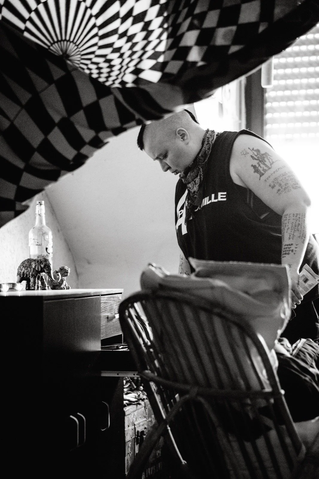 Schwarz-weiß-Fotografie eines Mannes mit Tattoos, der an einem Tisch arbeitet. Im Hintergrund sind eine Flasche, eine Elefantenfigur und Dekorationsgegenstände sichtbar. Der Raum ist gut beleuchtet durch ein Fenster mit Jalousien.