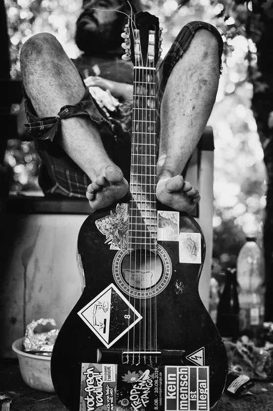 Ein Mann sitzt im Hintergrund mit einem akustischen Guitarre, die im Vordergrund steht. Die Gitarre ist mit mehreren Aufklebern bedeckt, darunter Warnhinweise und Logos. Der Mann trägt einen Bart und lange Haare, ist barfuß und hat die Beine hochgele