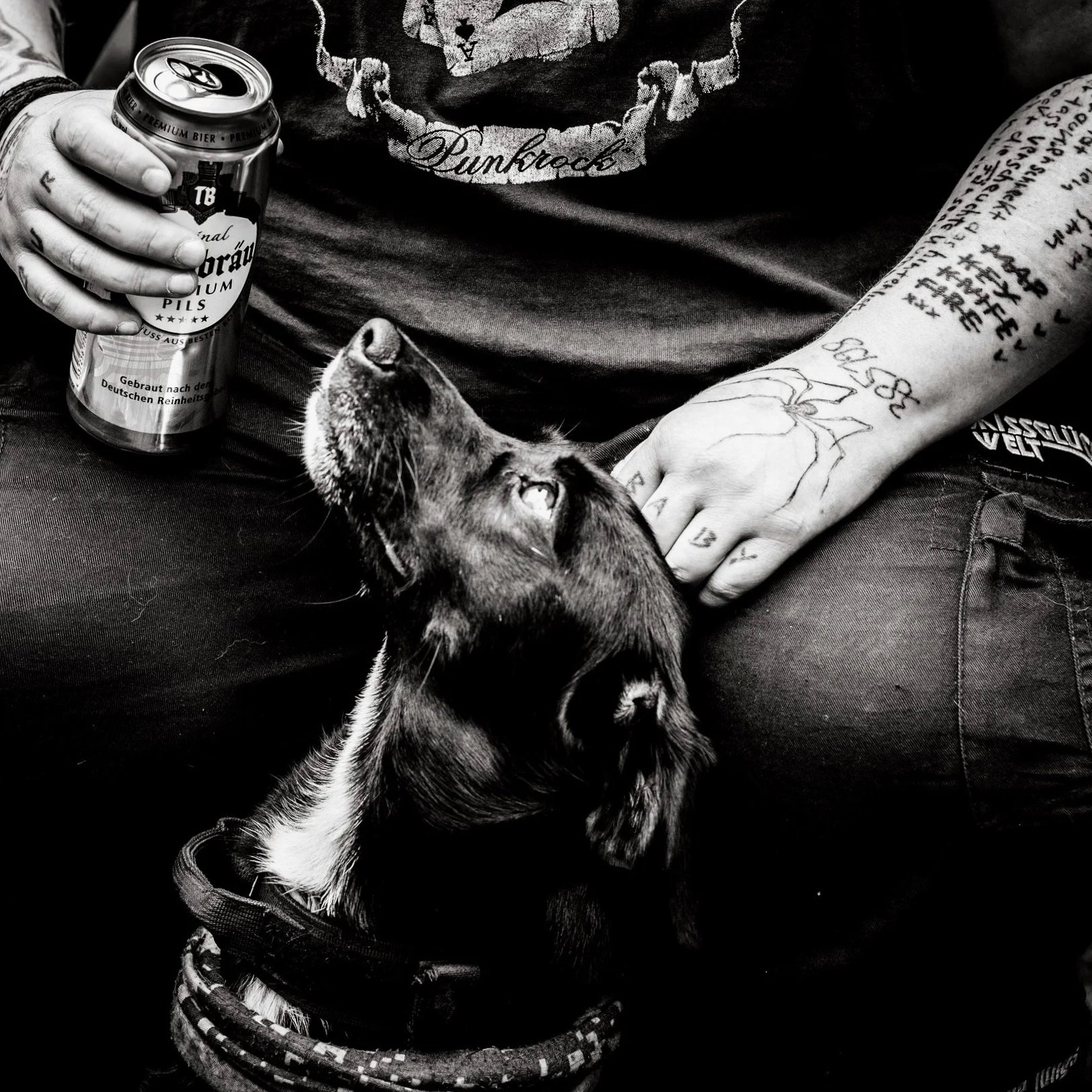 Nahaufnahme eines tätowierten Arms und einer Hand, die in die Kamera zeigt, alle in Schwarz-Weiß. Mehrere Tattoos sind sichtbar, inklusive Texten auf Armen und Hand, und es trägt einen Braided-Armband.