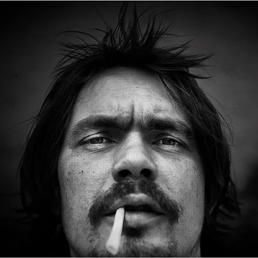 Nahaufnahme eines Mannes mit zerzausten Haaren, Schnurrbart und einem Zigarettenstummel im Mund, im Schwarz-Weiß-Foto.