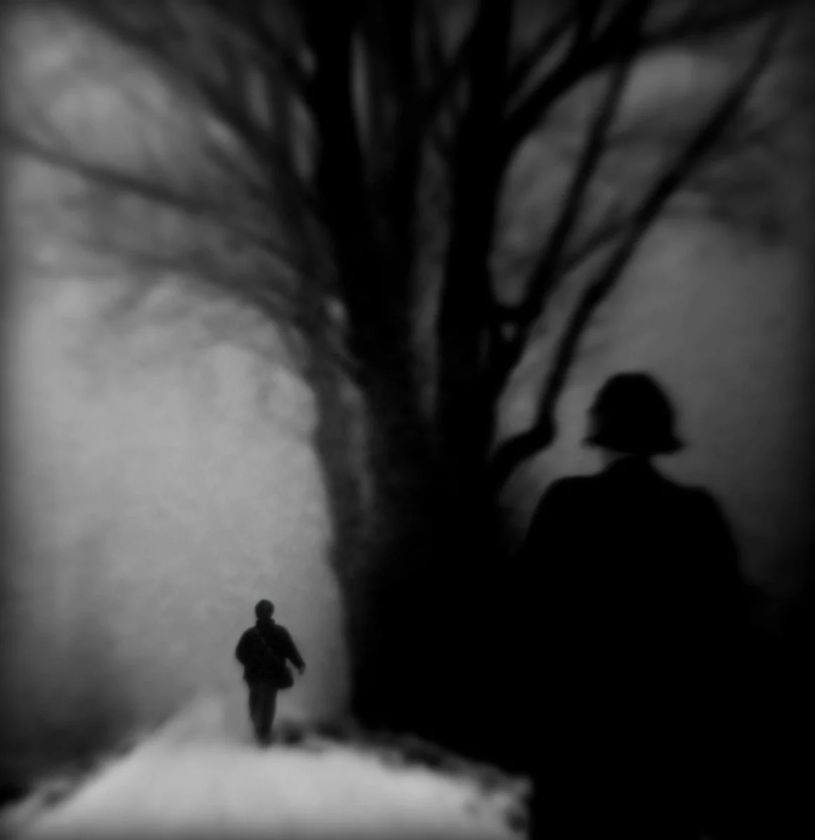 Schwarz-weiß-Foto eines Menschen, der auf einem verschneiten Weg geht, mit einem großen Baum im Hintergrund und einer großen menschlichen Silhouette im Schatten an der Seite.