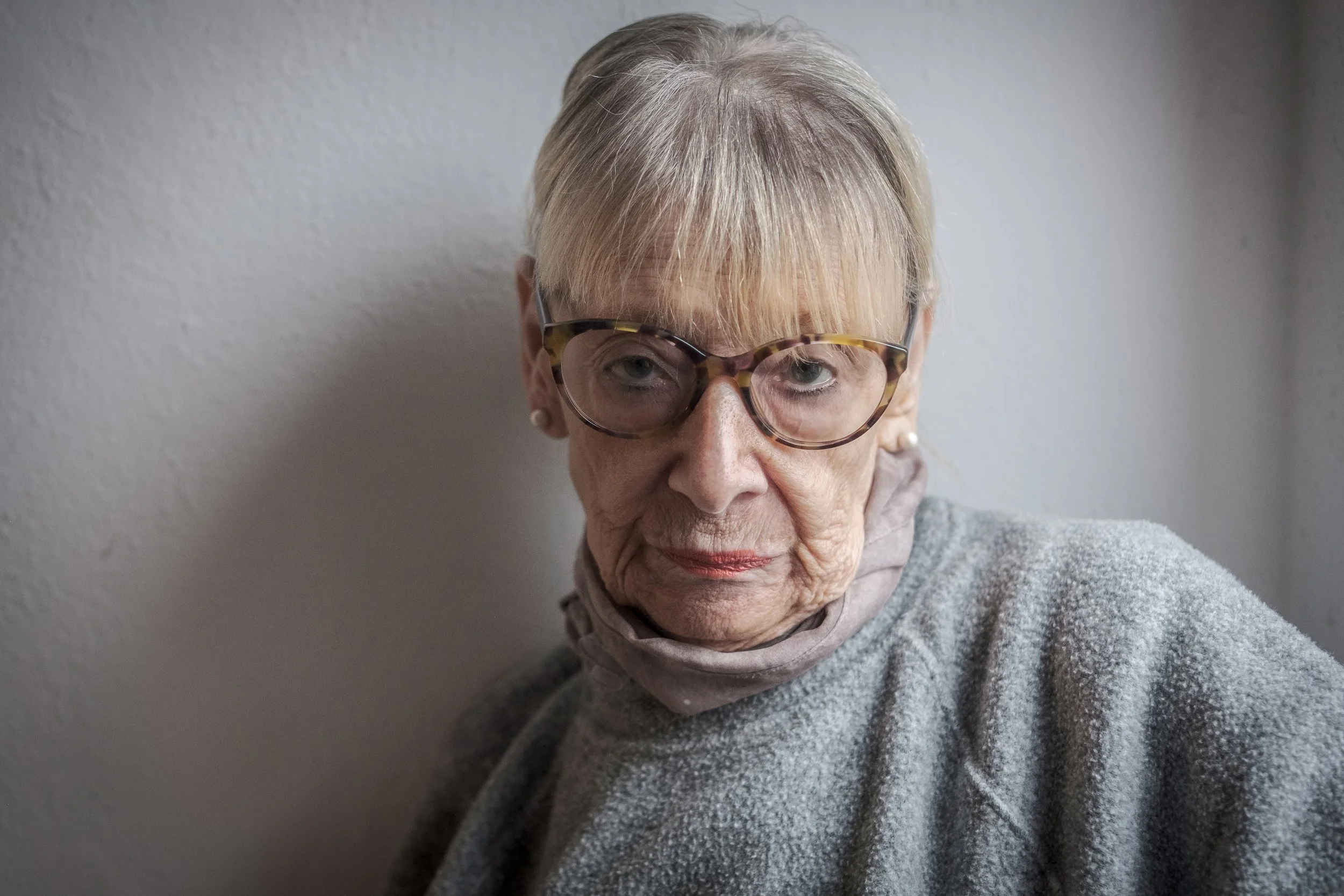 Nahaufnahme einer älteren Frau mit Brille und grauem Pullover vor einer weißen Wand.