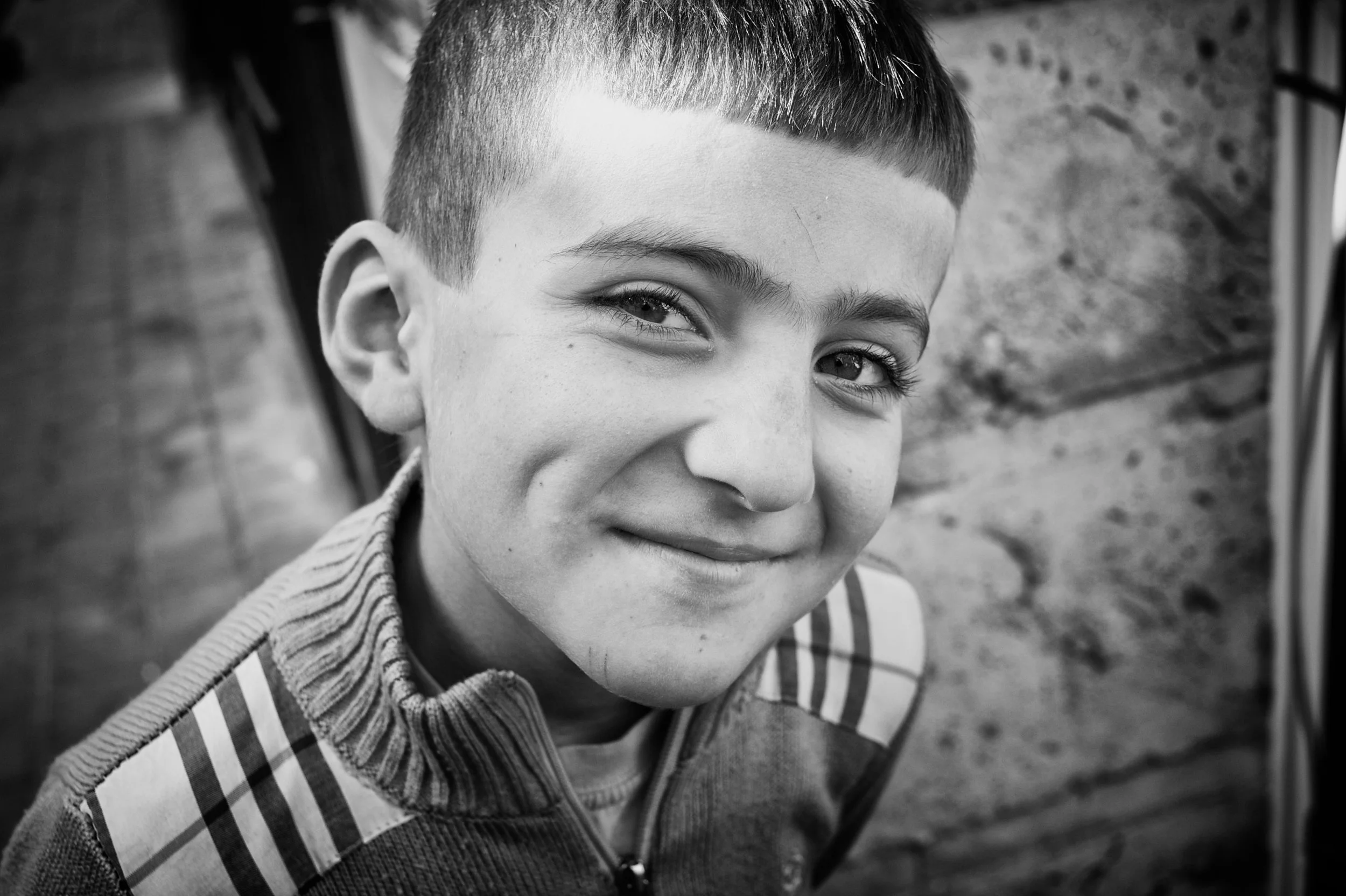 Nahaufnahme eines lachenden Jungen mit kurzen Haaren vor einer Steinmauer, in Schwarz-Weiß