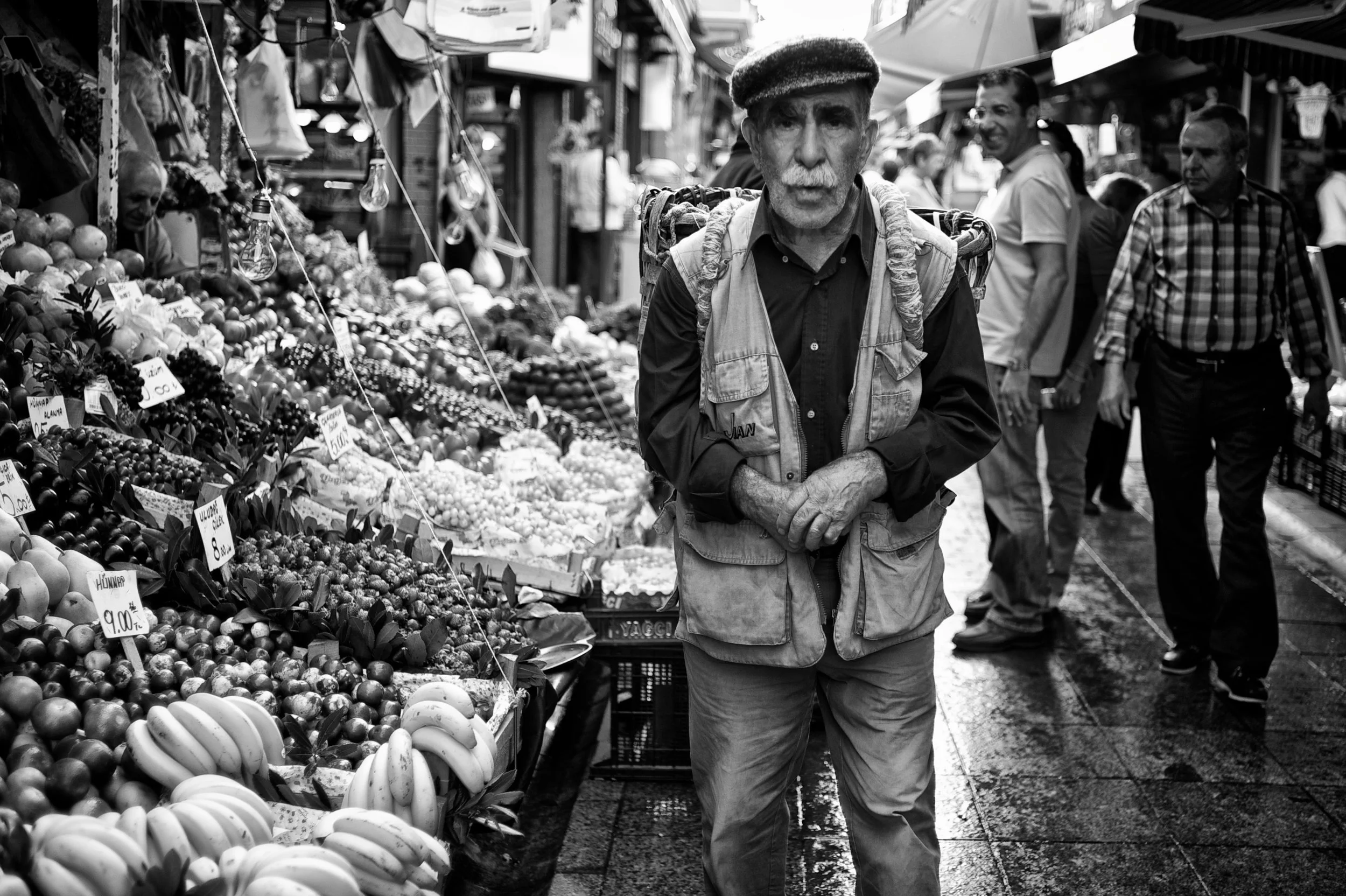 Ein älterer Mann mit weißem Schnurrbart und dunklem Hut steht auf einem belebten Markt, umgeben von Obstständen mit frischen Früchten. Er trägt eine Weste und schaut ernst in die Kamera.