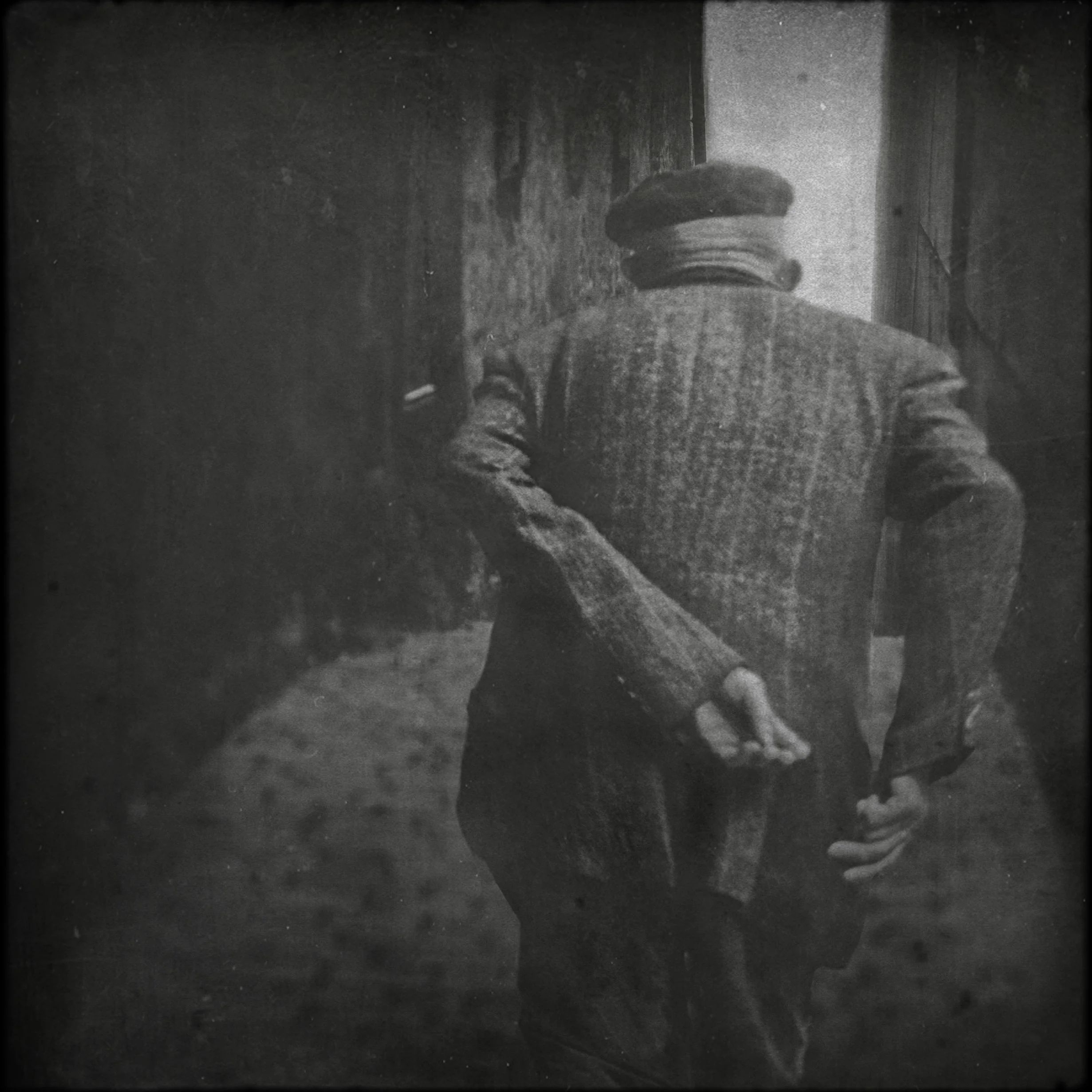 Schwarz-weiß-Fotografie eines Mannes von hinten, trägt einen Hut und einen Anzug, steht an einer Tür oder Mauer im Freien.