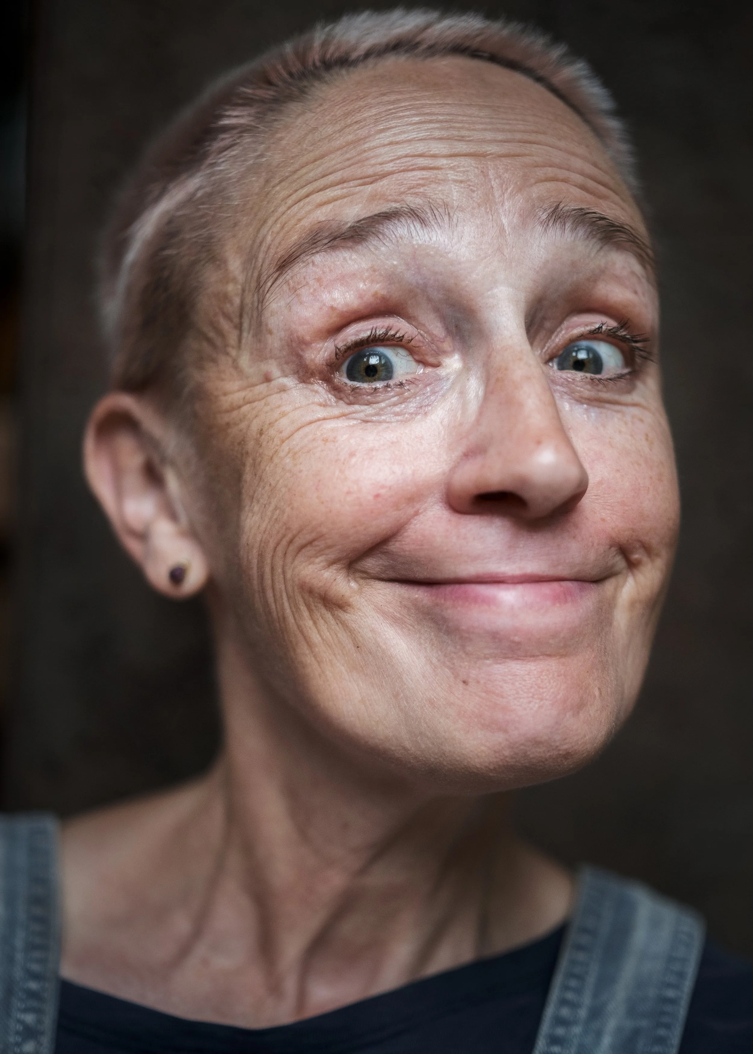 Nahaufnahme eines älteren lachenden Frauenkopfes mit kurzen, silbernen Haaren, blauen Augen, sichtbaren Falten und einem Ohrpiercing, vor dunklem Hintergrund.