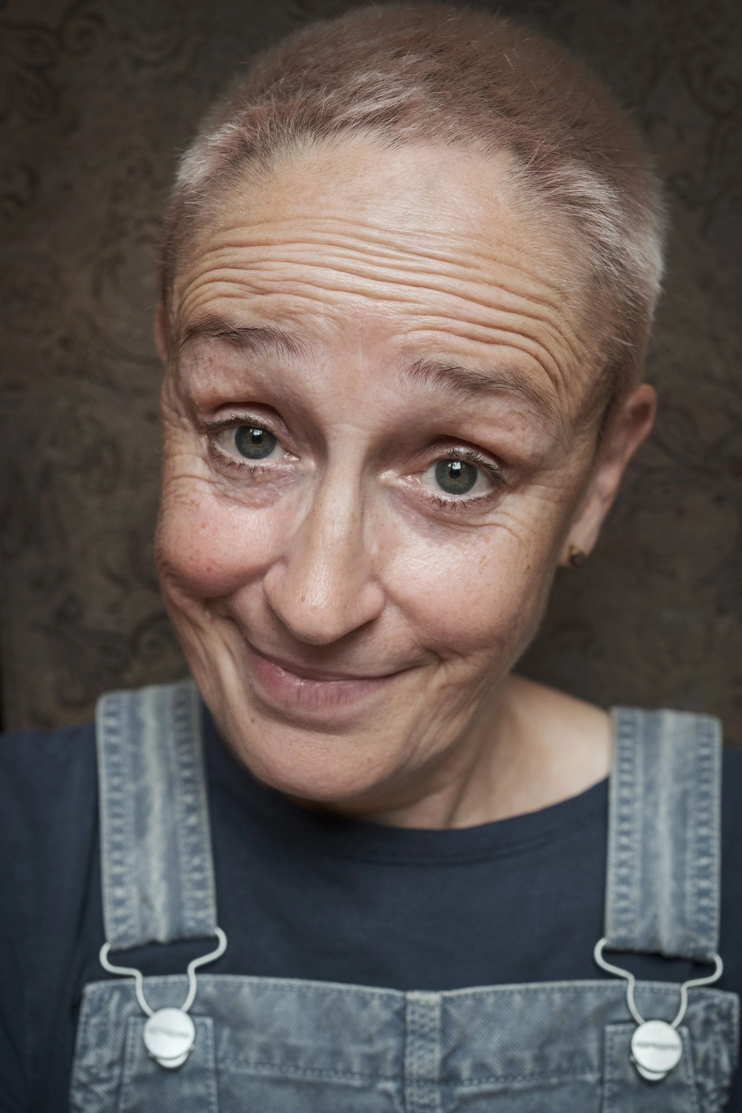 Nahaufnahme einer älteren Frau mit einem freundlichen Lächeln, grau-weißem kurzen Haar, trägt eine schwarze Oberseite und einen blauen Denim-Overall, vor einem dunklen Hintergrund.