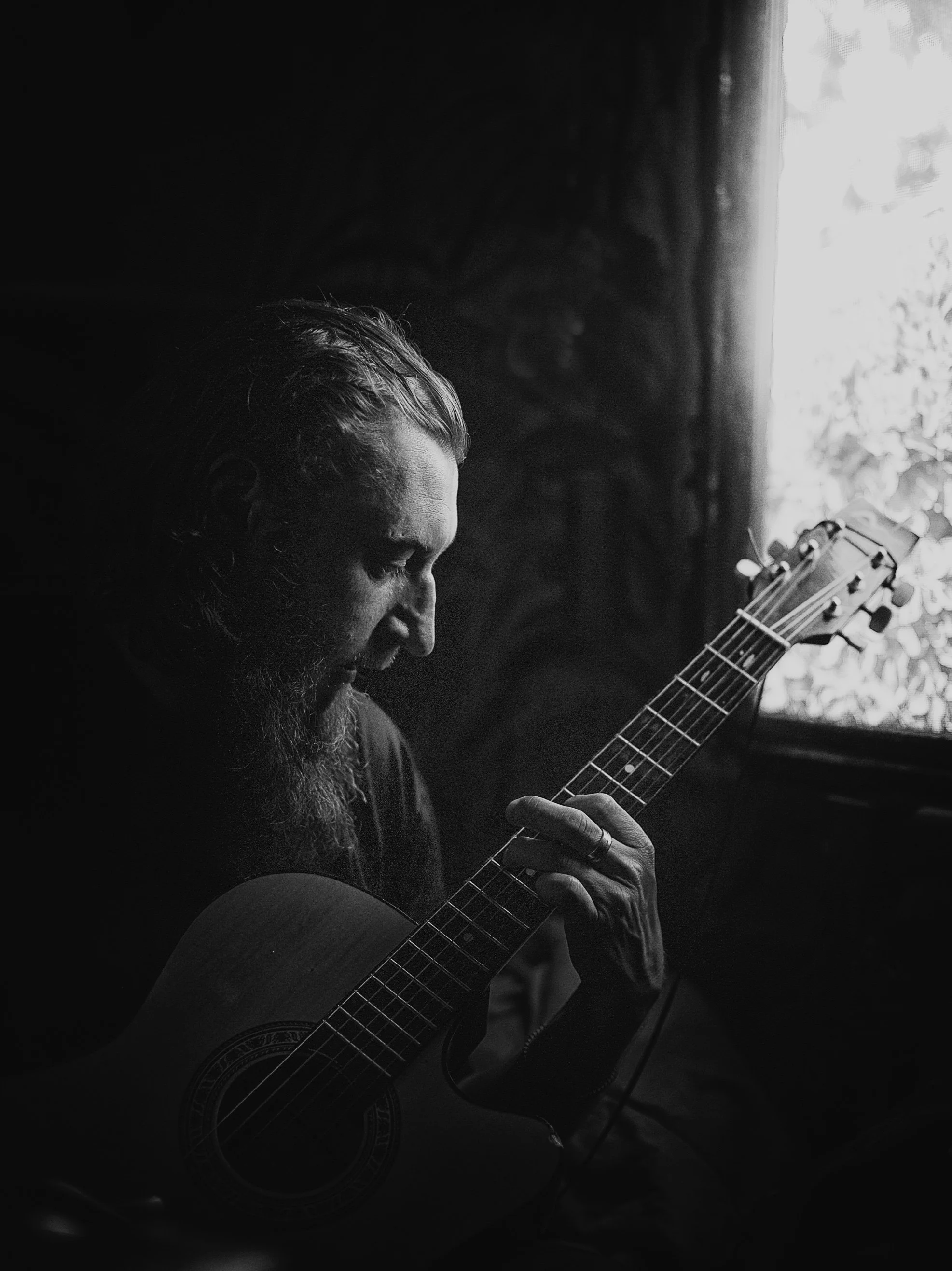 Schwarz-Weiß-Foto eines Mannes mit Vollbart, der Gitarre spielt, vor einer Fensteröffnung mit Licht. Der Mann ist seitlich zu sehen, die Stimmung ist ruhig und nachdenklich.