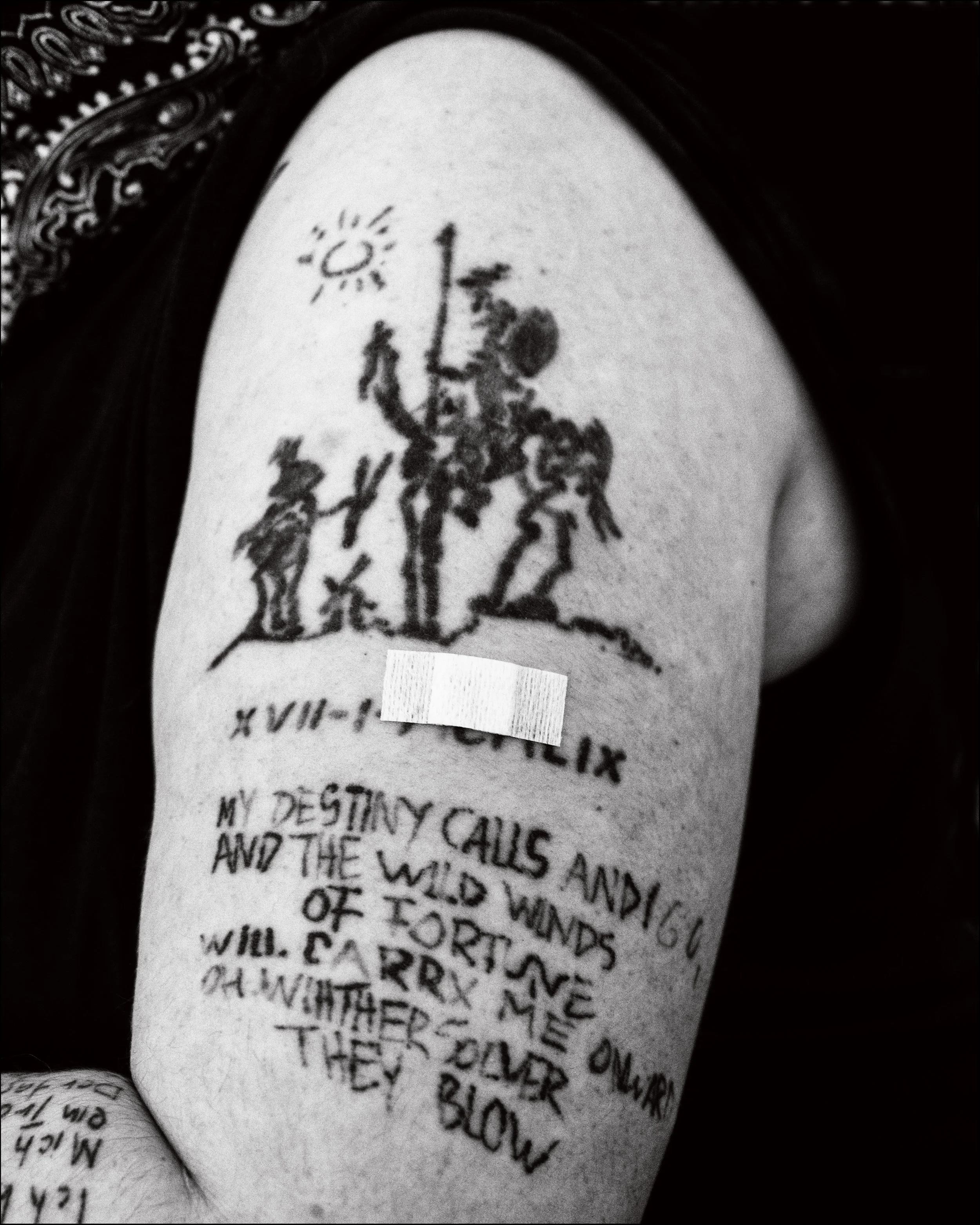 Schwarze Tätowierung auf dem Oberarm mit einer Silhouette von drei Figuren, einer mit einem Stab, und einem Sonnensymbol. Darunter ein verschnörkelter Text mit römischer Zahl X. Übersetzung des darunterstehenden Textes: 'My destiny calls and I go, an