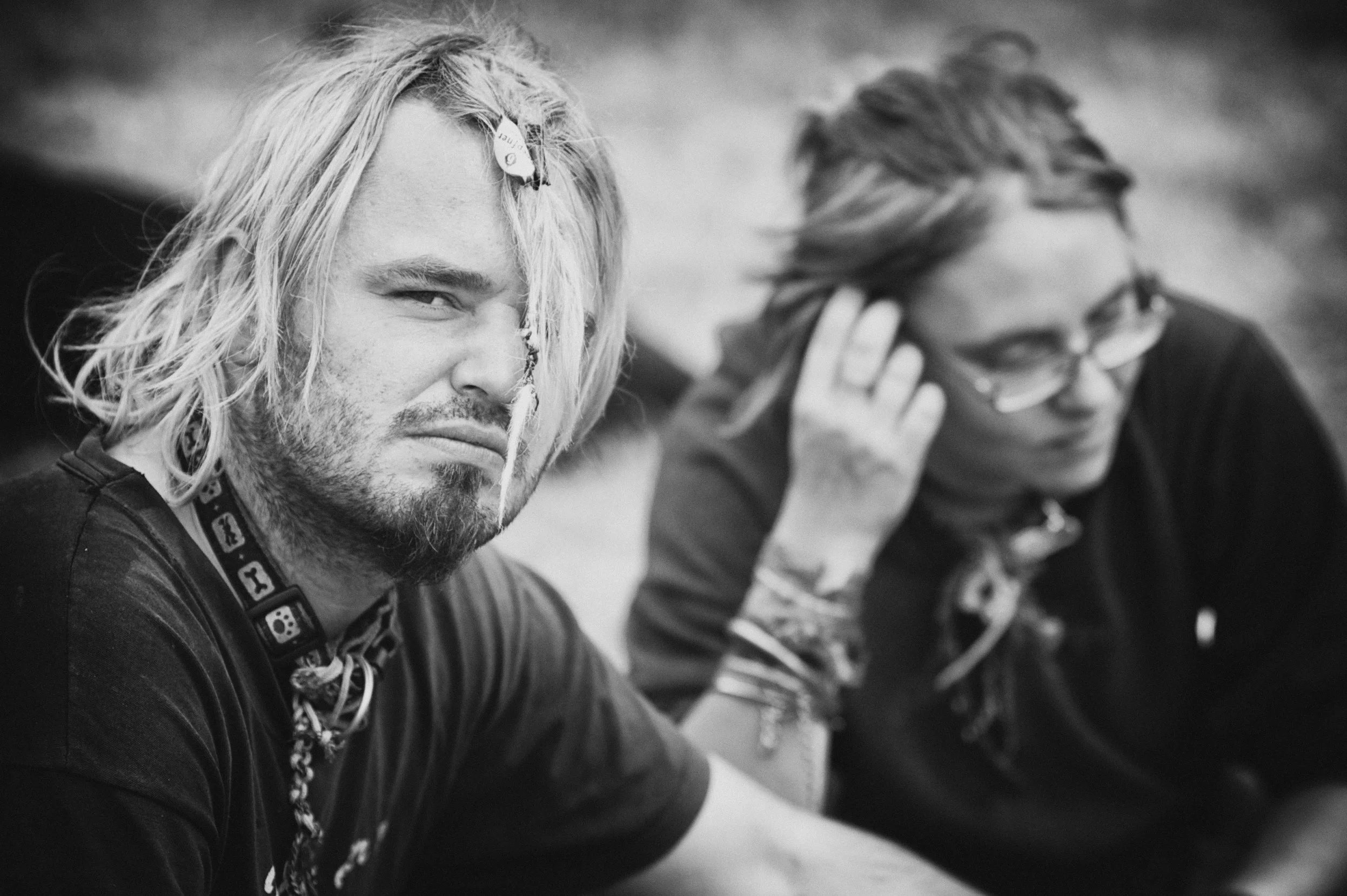 Zwei Menschen sitzen nebeneinander, derjenige im Vordergrund hat lange, lockige Haare, trägt eine Halskette mit Totenkopf und schaut ernst in die Kamera, die andere Person trägt Brille, hat kürzere Haare und lächelt leicht, während sie an ihrem Ohr w