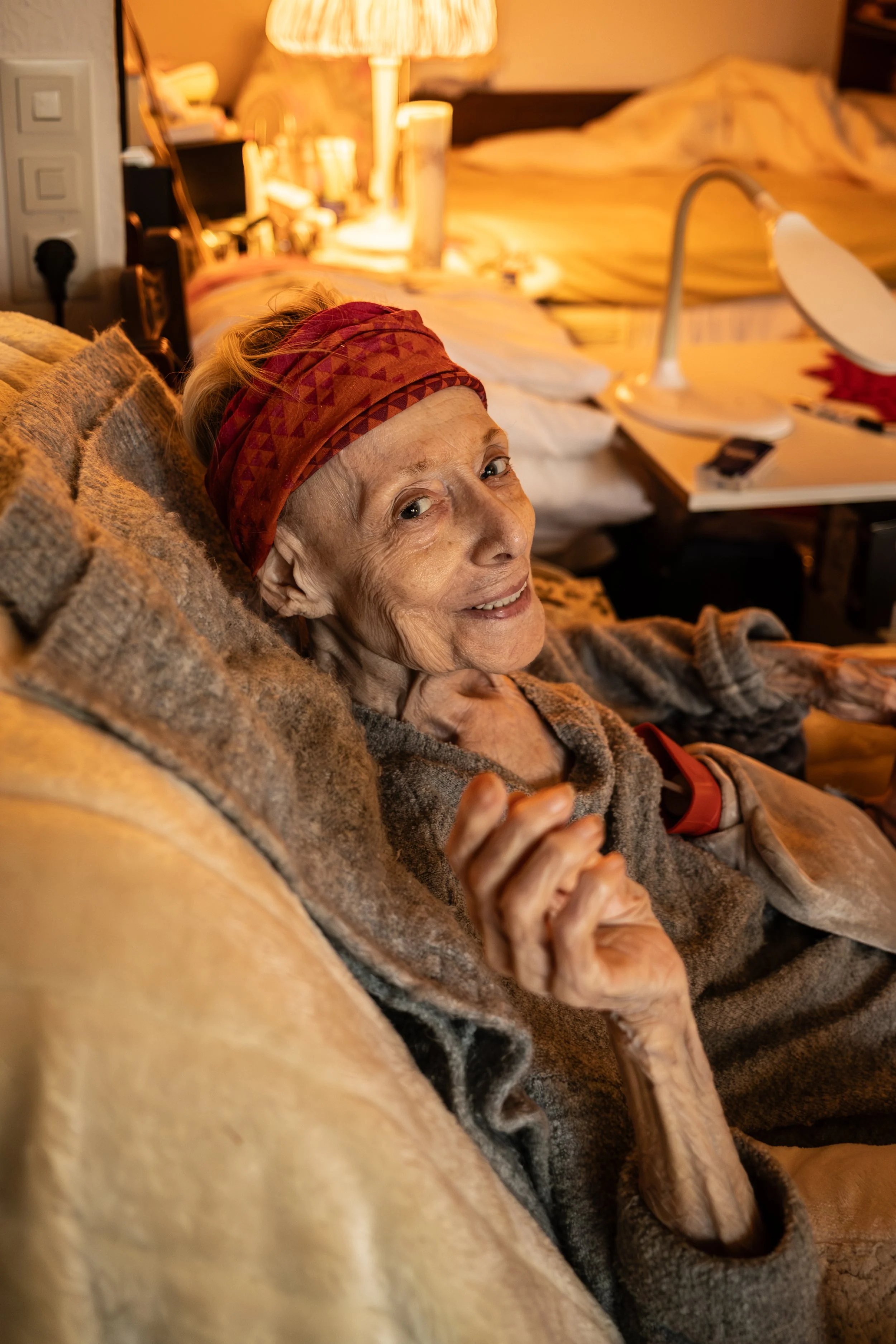 Eine ältere Frau liegt entspannt auf einer Couch in einem gemütlichen Raum, trägt einen roten Stirnband und lächelt in die Kamera.