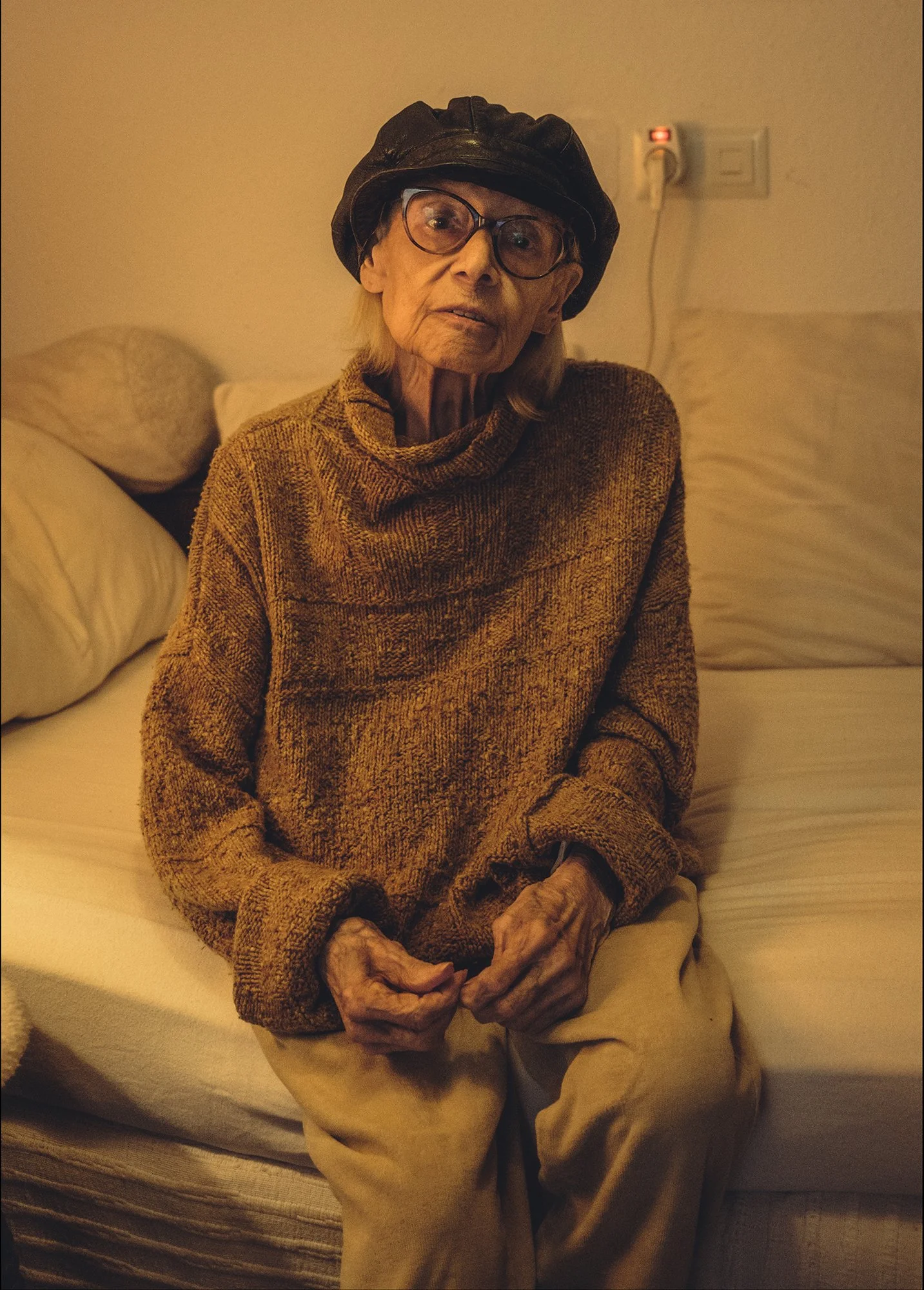 Ältere Frau sitzt auf einem Bett, trägt eine braune Strickjacke, eine schwarze Mütze und große Brille, vor einer beige Wand mit Stromanschluss.