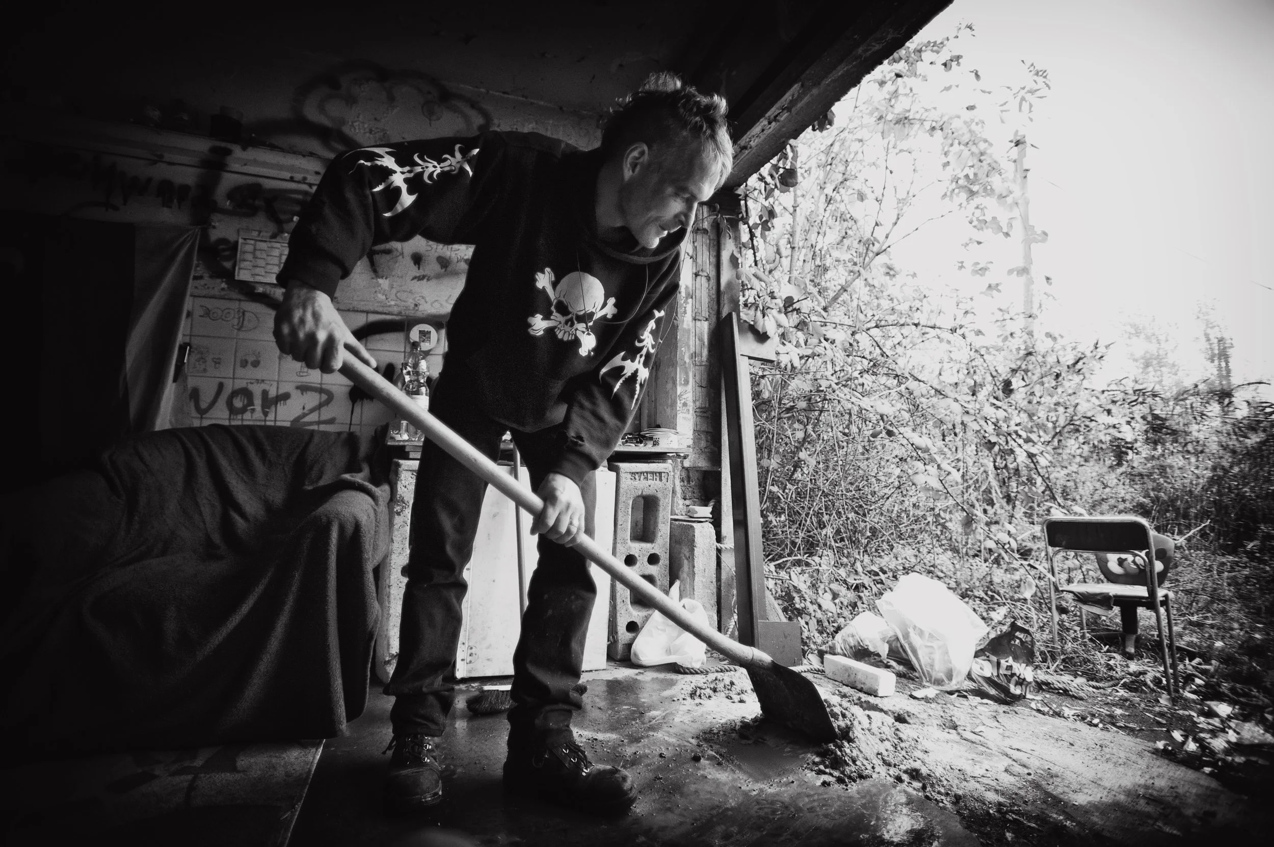 Mann beim Schaufeln von Erde oder Sand in einem dunklen Raum mit offenem Eingang nach draußen, wo Bäume und ein Stuhl sichtbar sind. Der Mann trägt ein T-Shirt mit Totenkopf-Design.