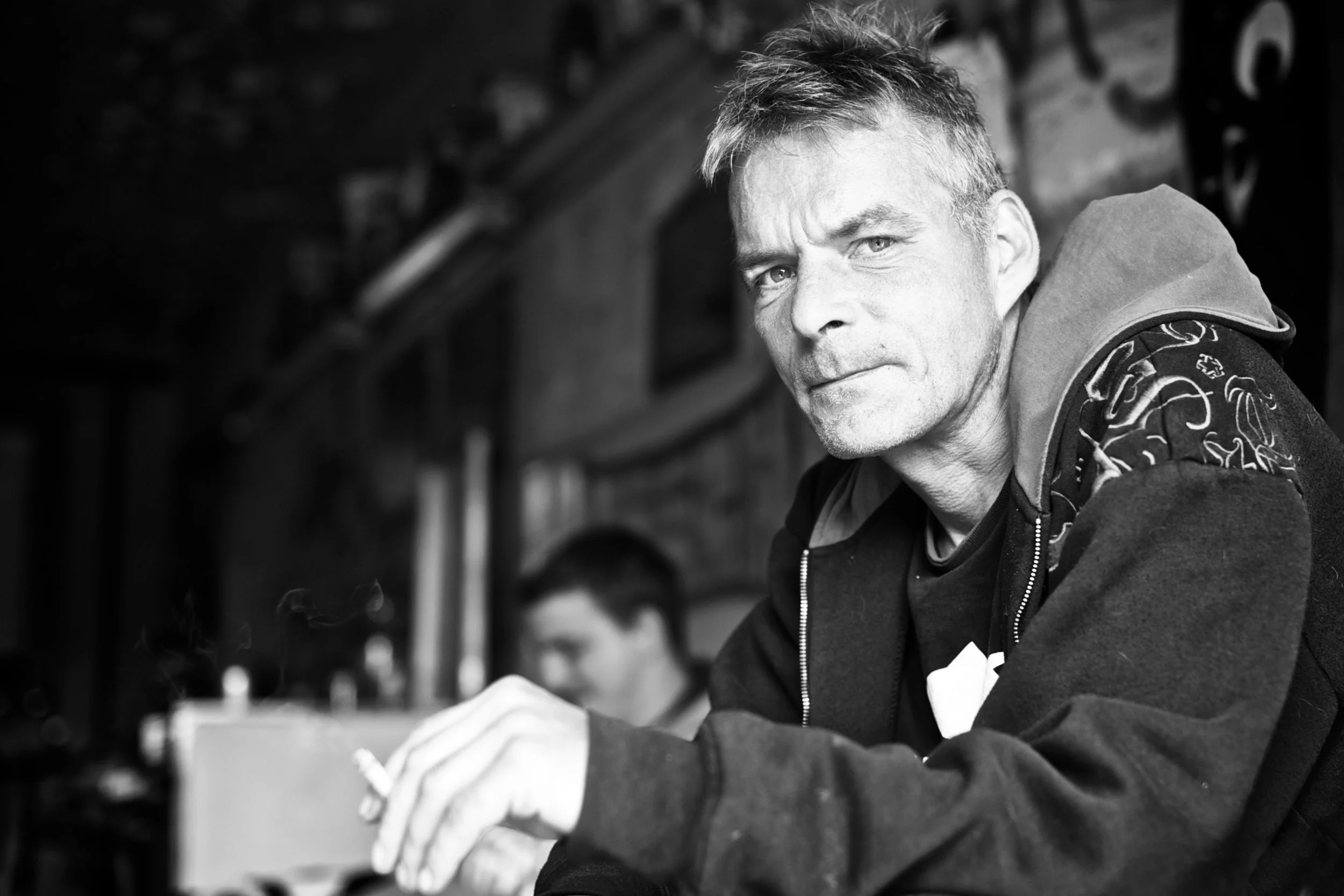 Schwarz-Weiß-Fotografie eines Mannes mit kurzen, grauen Haaren, der in die Kamera schaut. Er trägt eine dunkle Jacke mit einem grafischen Muster auf den Schultern und eine Kapuze. Im Hintergrund ist eine zweite Person zu sehen, die unscharf ist