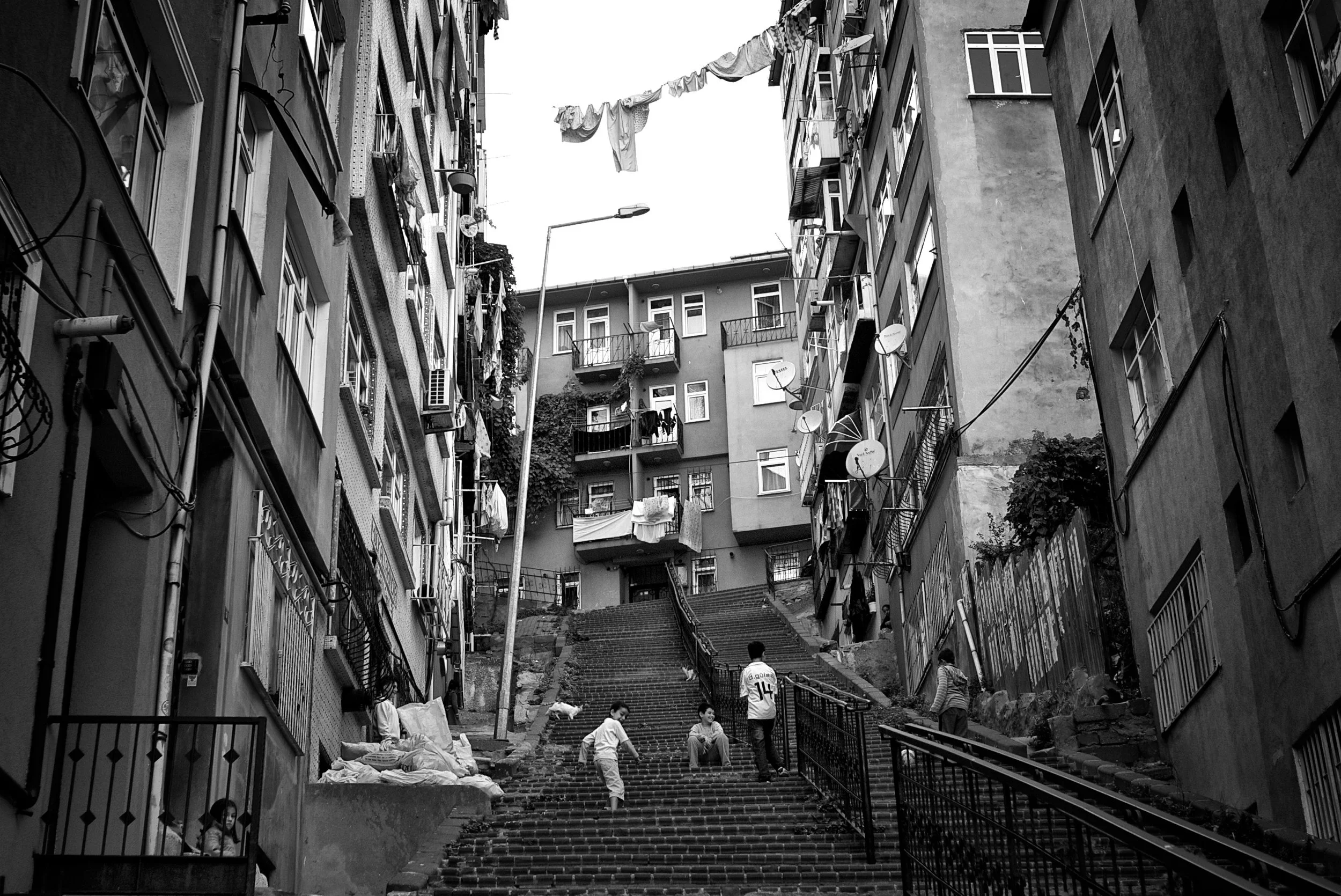Schwarz-weiß-Foto von einer steilen Treppe zwischen hohen Apartmentgebäuden mit Wäsche und Satellitenschüsseln an den Balkonen, Kinder spielen auf den Stufen, im Hintergrund Wohnhäuser.