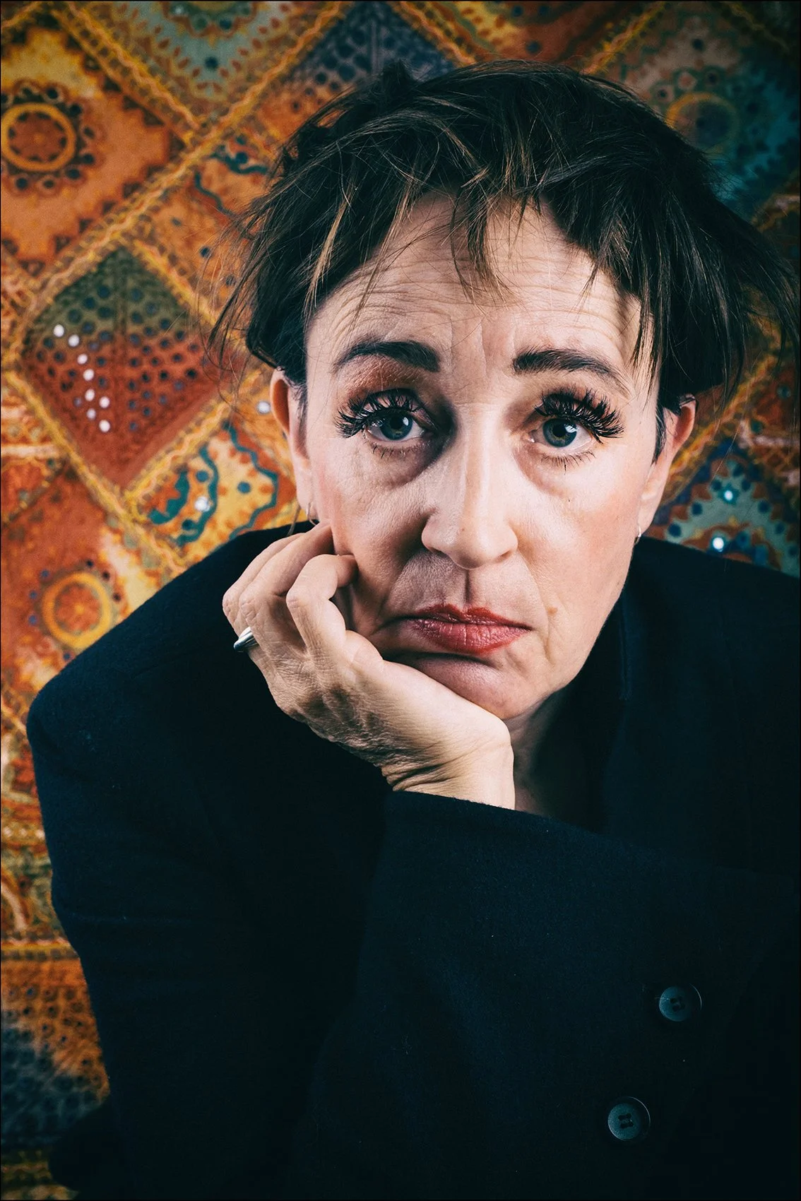 Nahaufnahme einer Frau mit dunklem, zerzaustem Haar, die auf einem bunten, orientalisch inspirierten Teppich sitzt. Sie trägt einen schwarzen Mantel und schaut direkt in die Kamera, die Hand stützt ihr Gesicht.