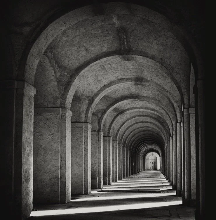 Blick durch einen langen, archtikierten Flur mit wiederholten Bögen und Schatten im Schwarz-Weiß-Design.