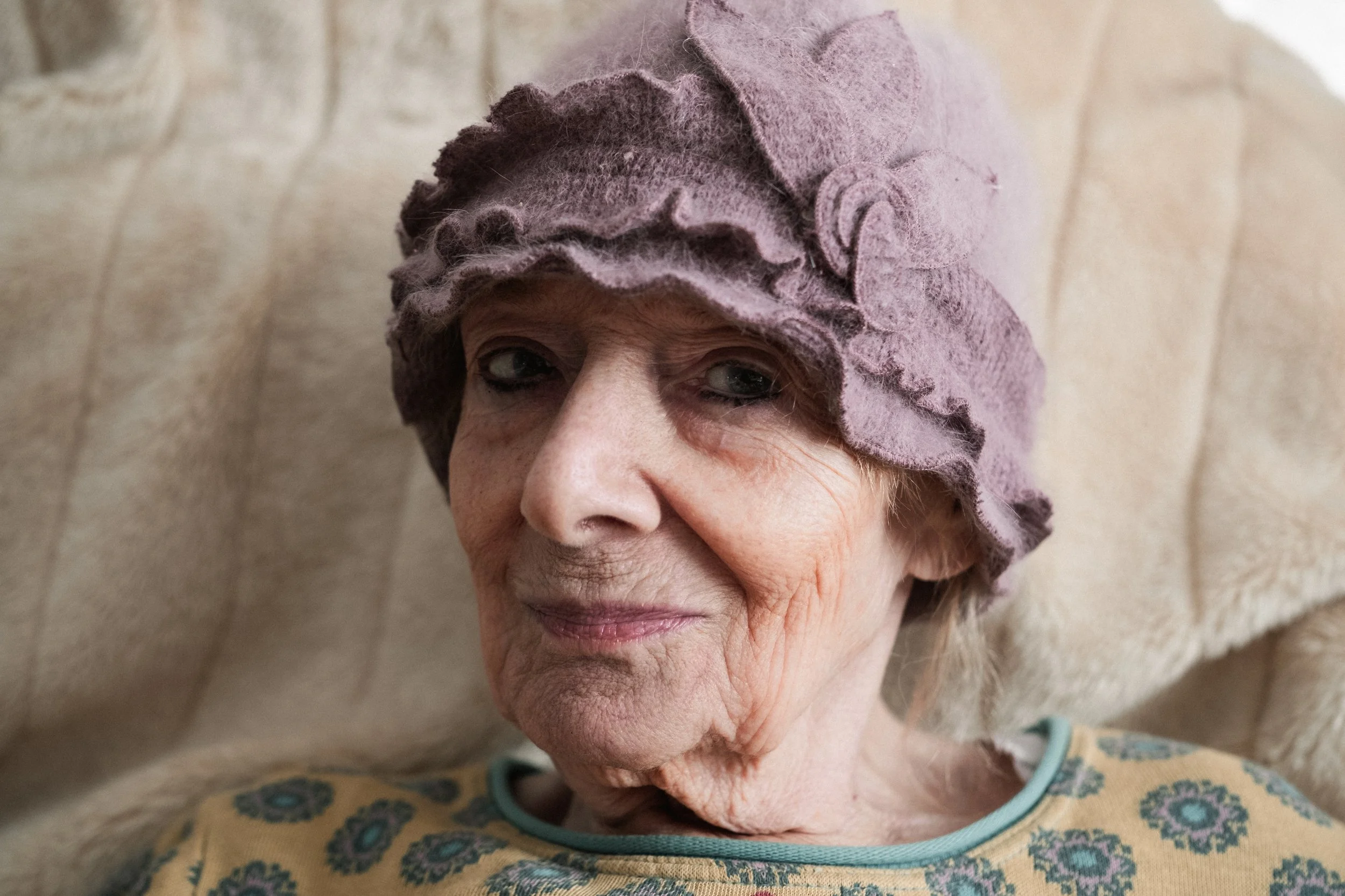 Ältere Frau mit lila Hut, sitzt auf einem hellen, gesteppten Sofa, trägt ein gelbes Oberteil mit Blumenmuster.