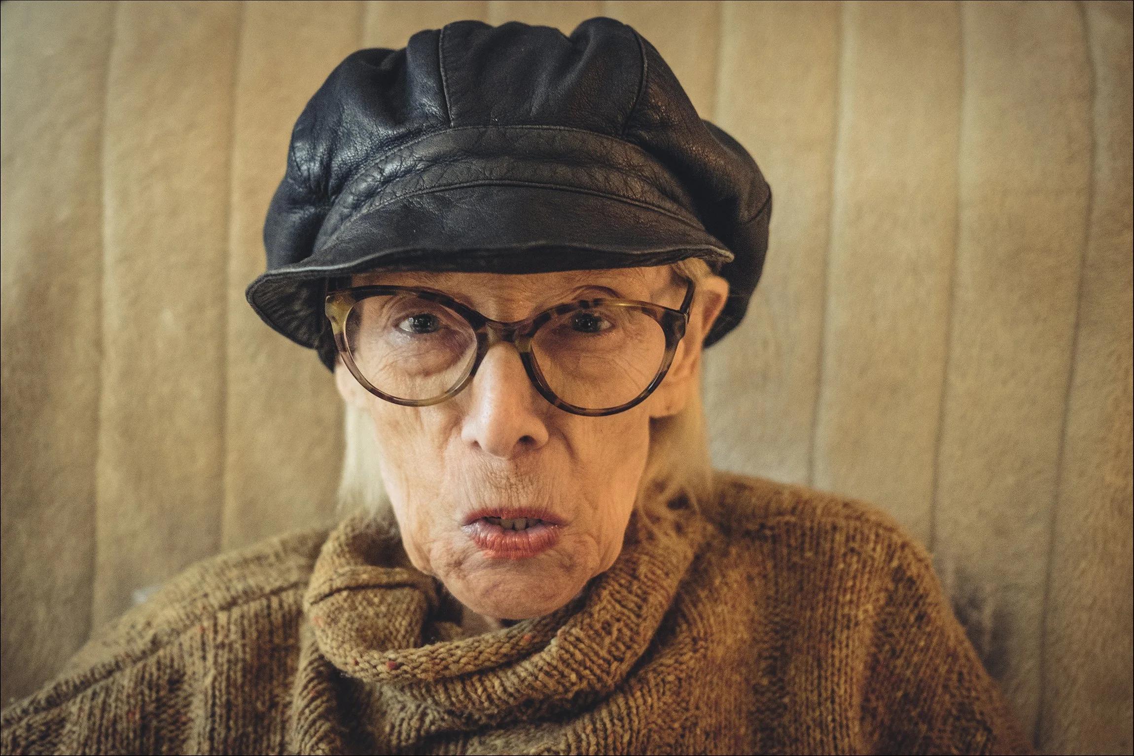 Nahaufnahme einer alten Frau mit Brille, Lederkappe und braunem Pullover, die auf einem Sofa sitzt.