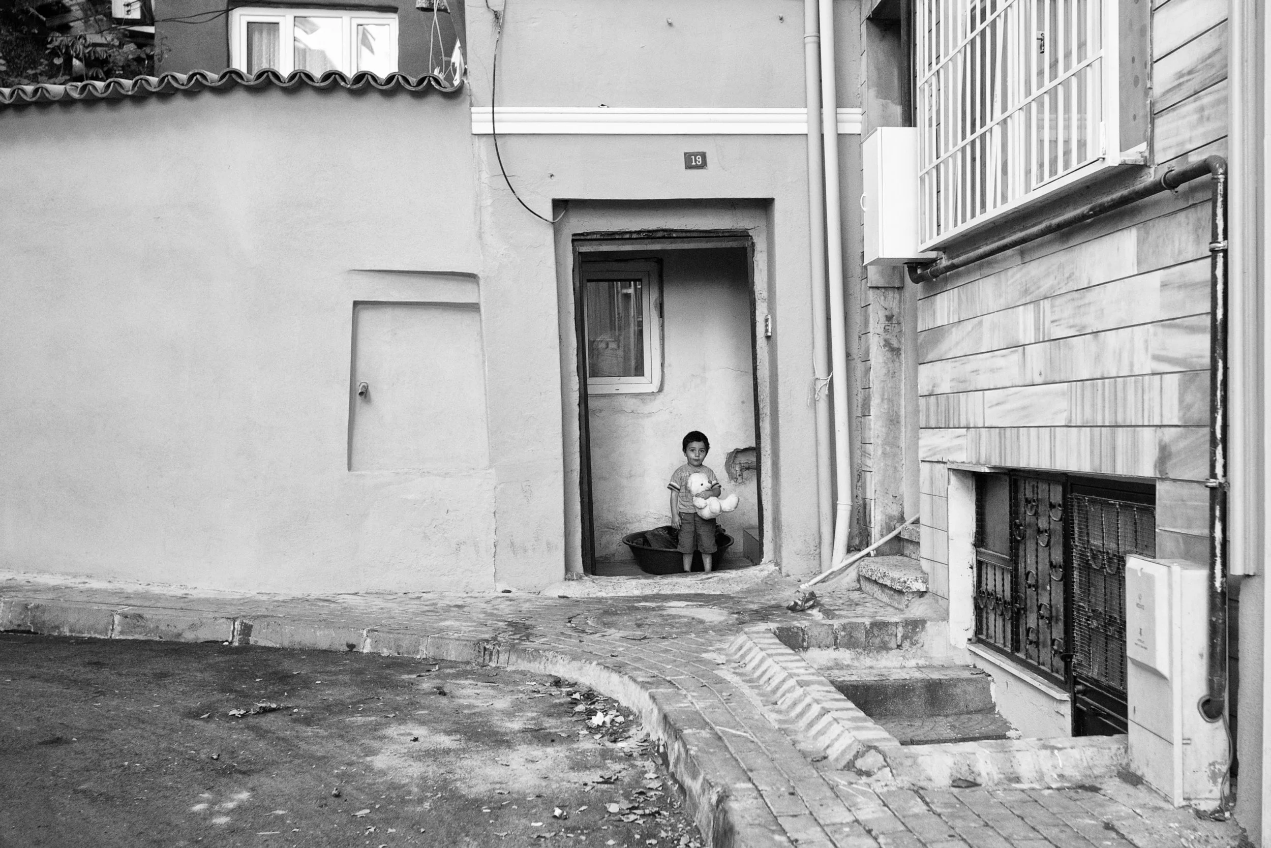 Ein Junge steht in einer Türöffnung vor einem Haus, hält ein Plüschtier und blickt nach vorne. Das Bild ist schwarz-weiß.