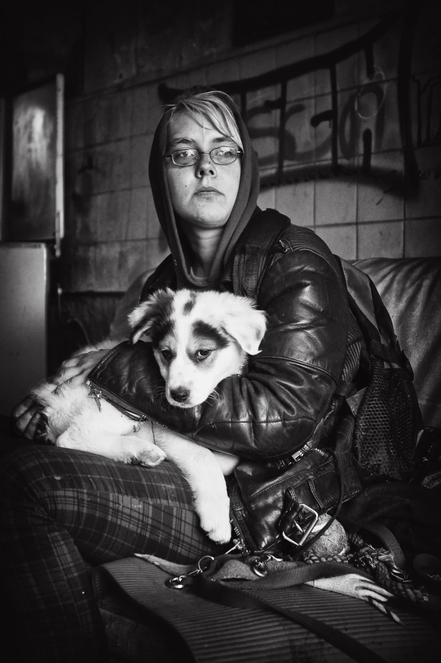 Schwarz-weisses Foto einer jungen Frau mit kurzen Haaren und Brille, die einen Welpen auf ihrem Schoß hält, in einem urbanen, grauen Umfeld mit Graffiti an der Wand.