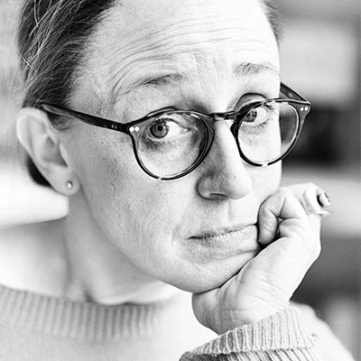 Nahaufnahme einer älteren Frau mit kurzen Haaren, Brille und nachdenklichem Ausdruck, die ihre Hand an ihr Gesicht legt.