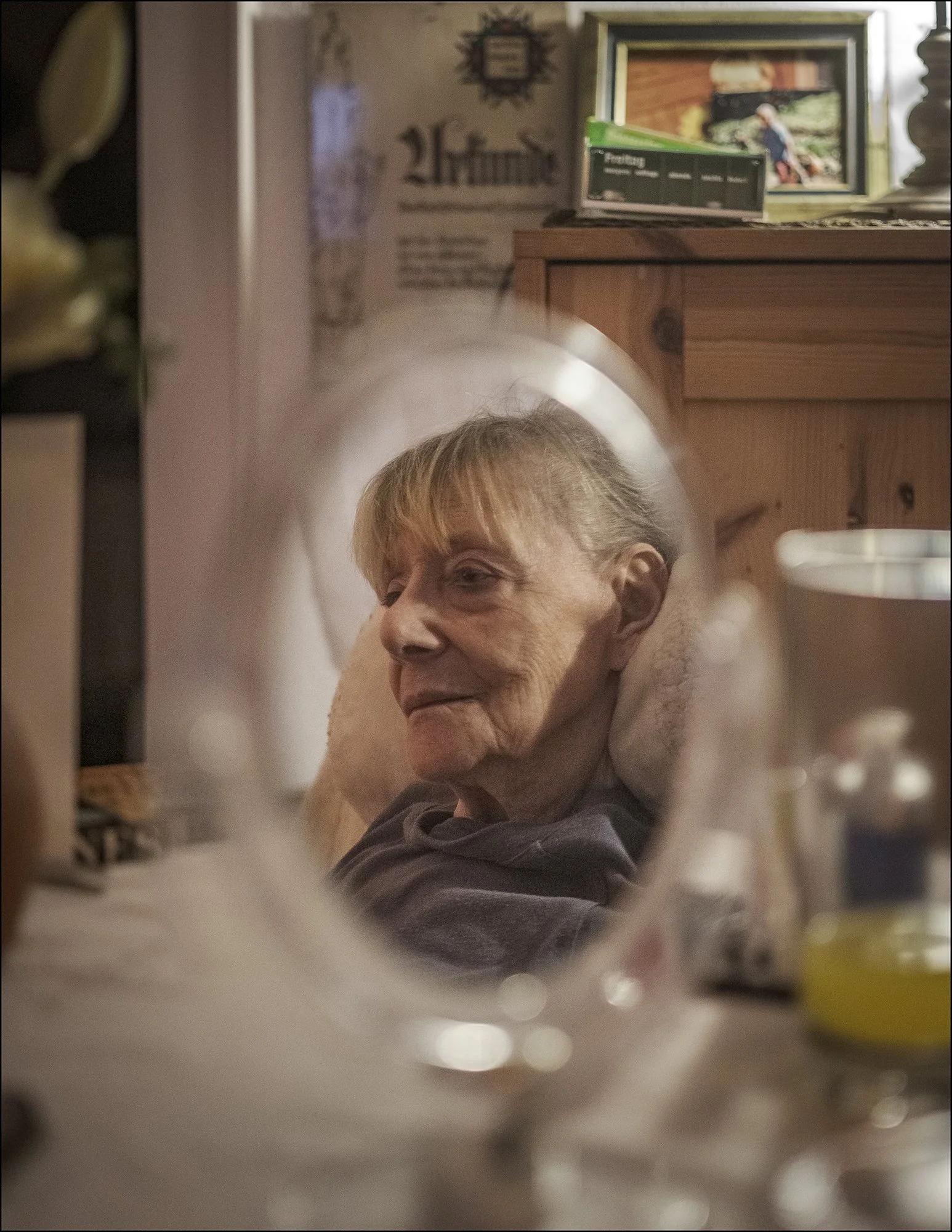 Eine alte Frau sitzt entspannt auf einem Sofa, ihr Blick ist nach vorne gerichtet, im Vordergrund ist ein Spiegel, der ihr Bild reflektiert, und im Hintergrund sind Bilder und Dekorationen an der Wand zu sehen.