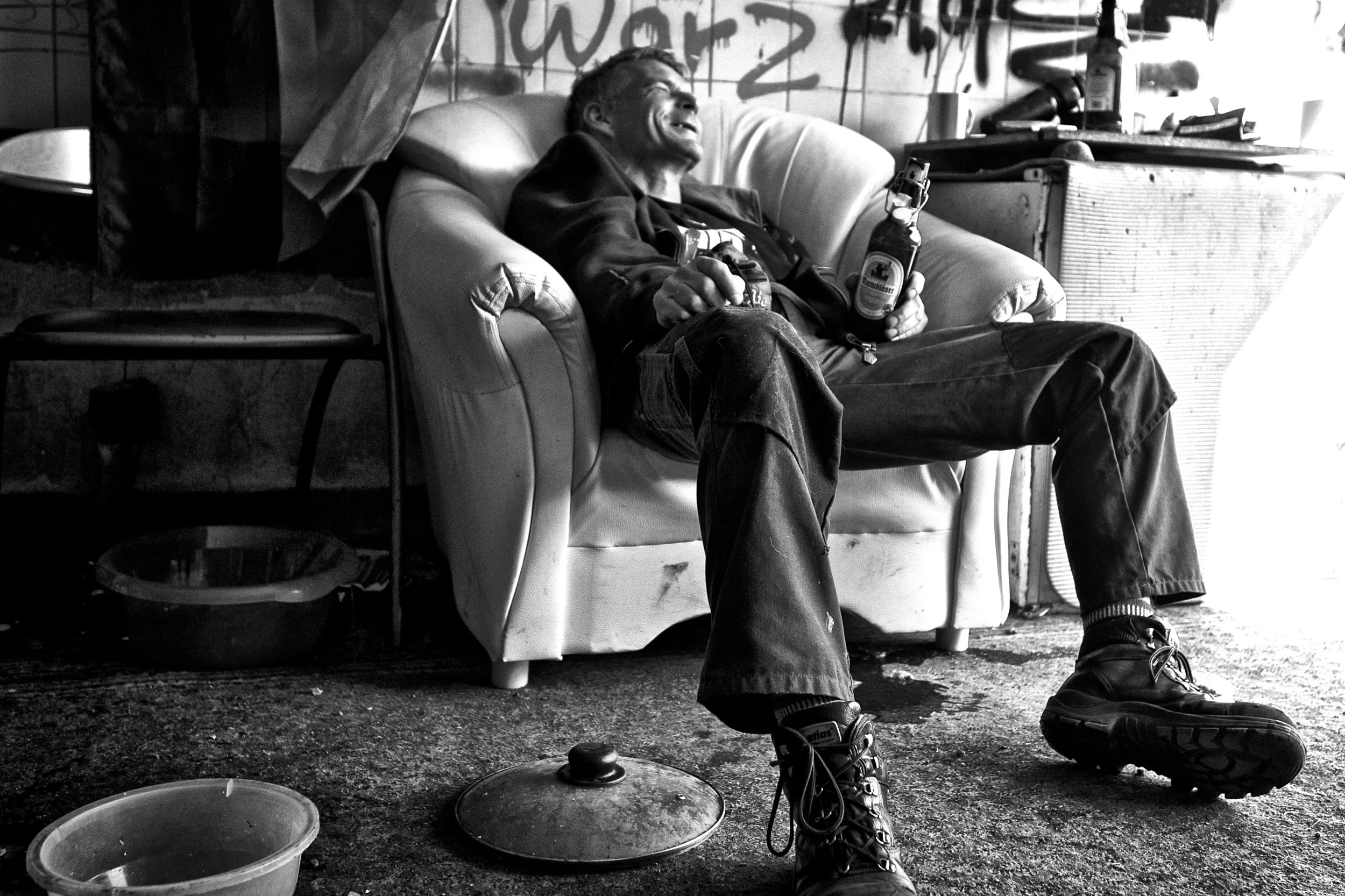 Ein Mann sitzt auf einem Sofa, trinkt Bier und lacht in einem schmutzigen Raum.