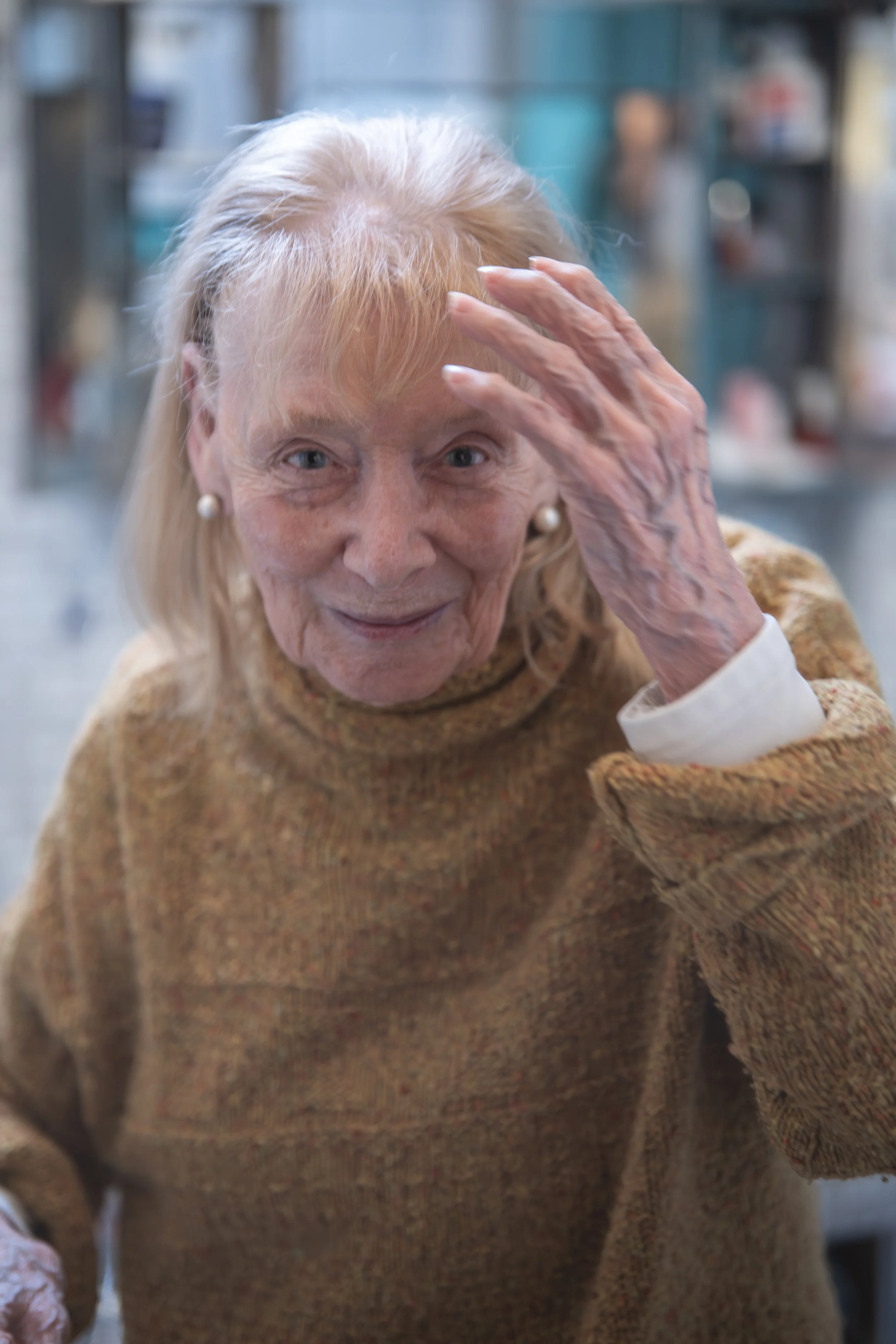 Eine ältere Frau mit blondem Haar, trug einen braunen Pullover und Edelstecker, lächelnd und die Hand auf der Stirn, in einem Raum mit Bücherregalen im Hintergrund.