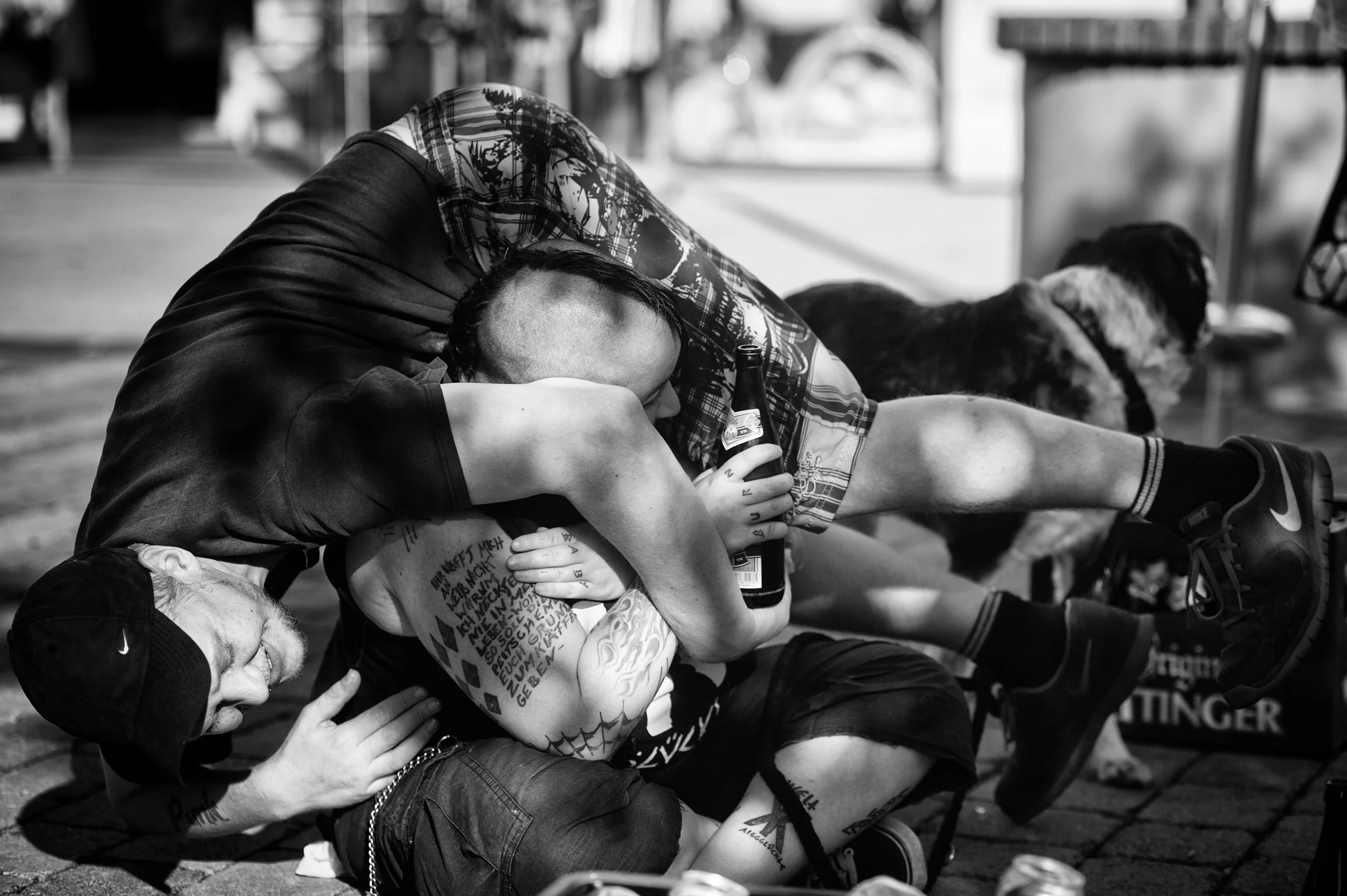 Zwei Männer im Kampf auf der Straße, einer liegt auf dem Boden, der andere ist in einer Übergriffposition, während sie eine Flasche halten. Es ist eine schwarze und weiße Fotografie.