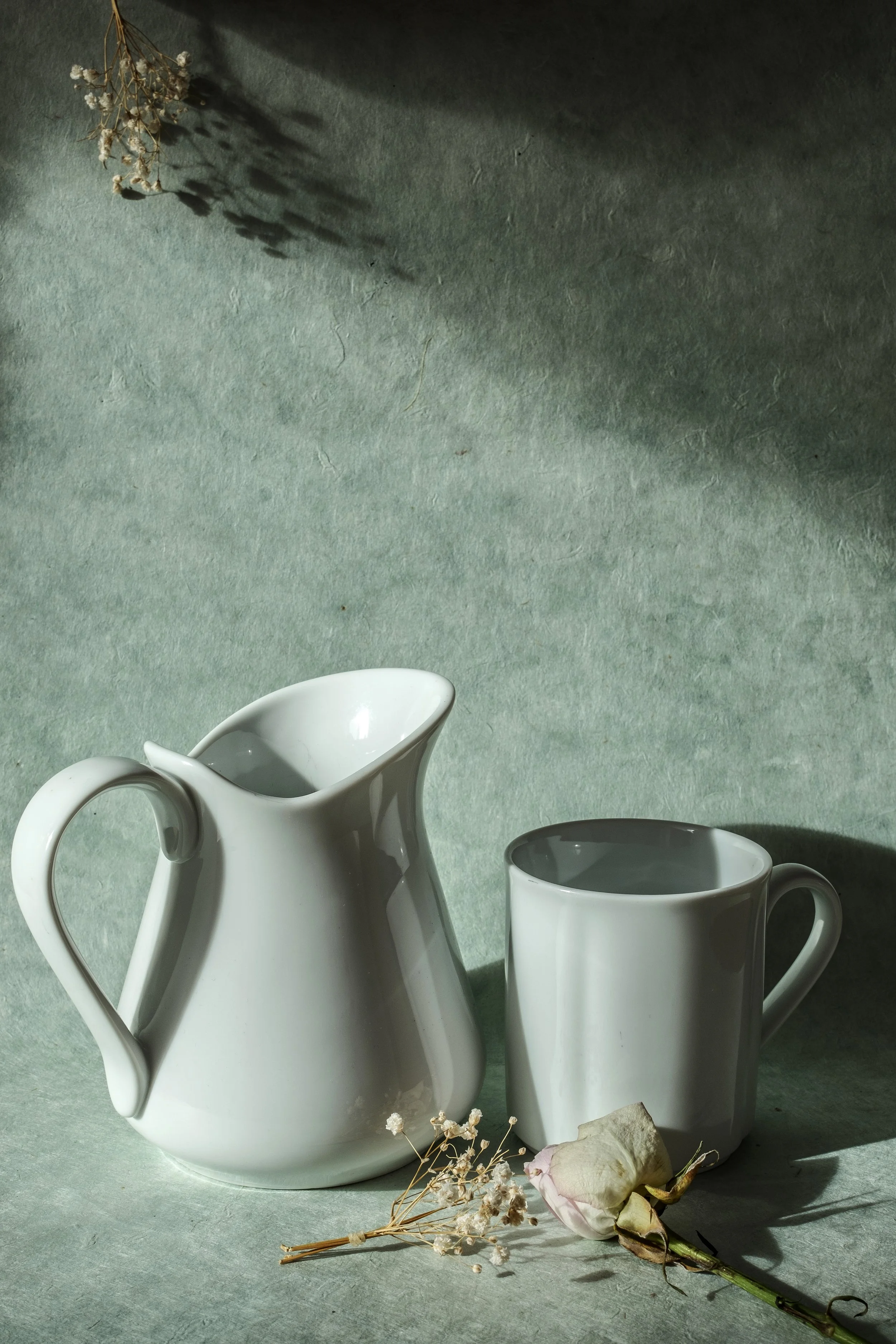Weiße Keramik-Kanne und Tasse, trockene weiße Rose und kleine Blumen, Hintergrund aus Papier in Grau, mit Schatteneffekt.