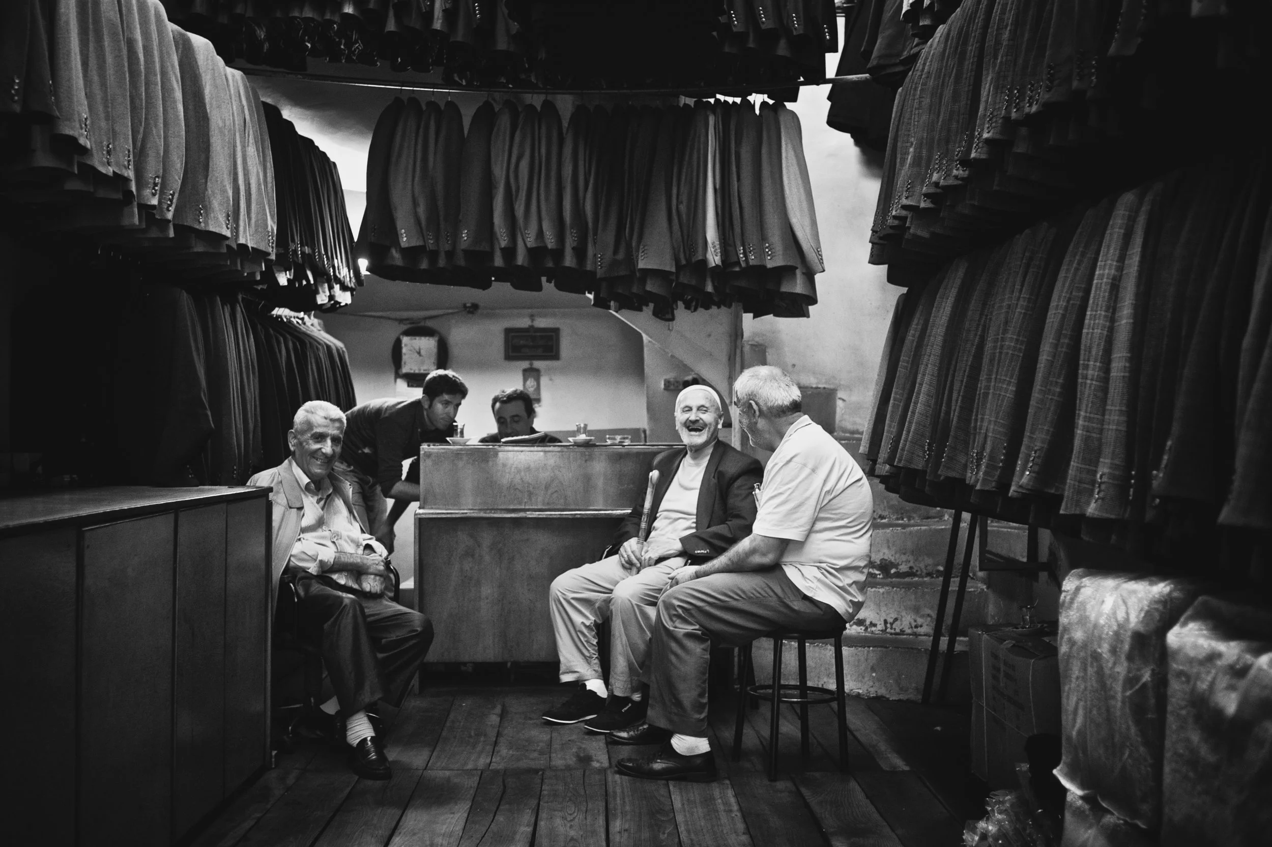 Fünf Männer in einem Bekleidungsgeschäft, einige sitzen und lachen, während andere im Hintergrund stehen und schauen, alle in schwarz-weiß.