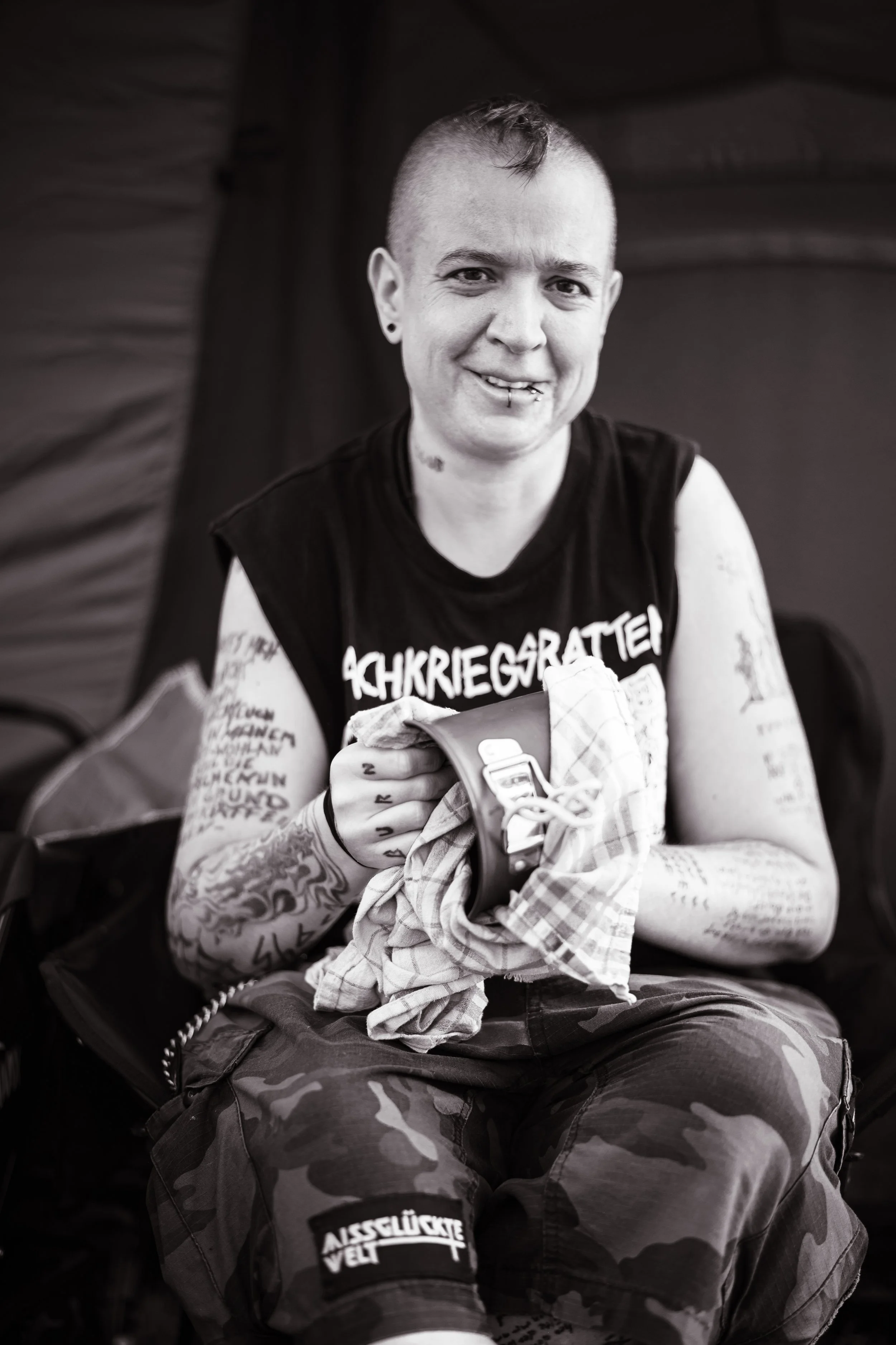Person mit kurzen, rasierten Haaren, wachsendem Rücken, tätowierten Armen, lächelnd, in Camouflage-Hose, im schwarzen T-Shirt mit Aufdruck, sitzt und hält ein Tuch und einen Gegenstand in den Händen.