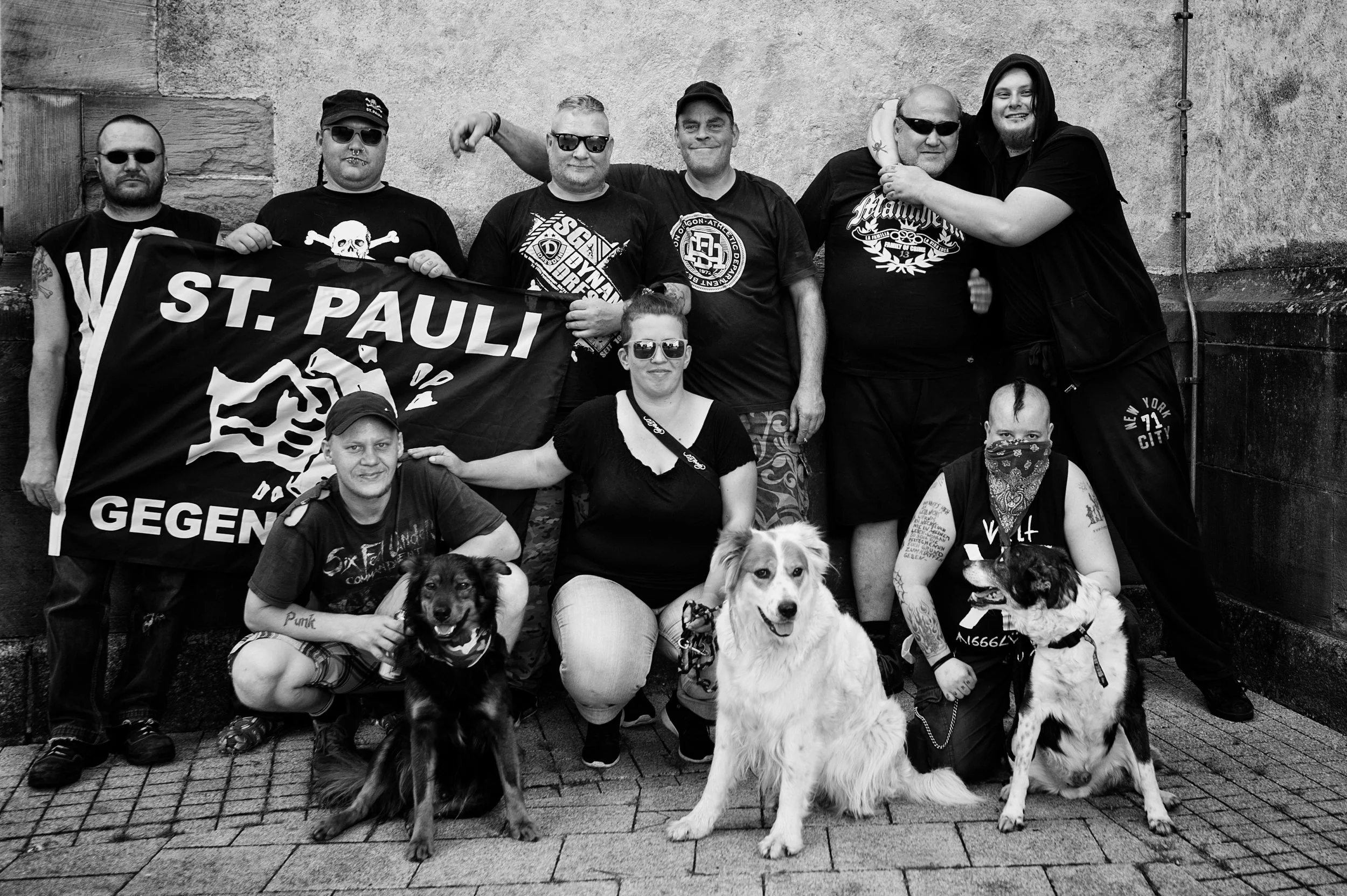 Gruppe von Menschen mit Hunden posiert vor einer Wand, einige tragen Sonnenbrillen, eine Person trägt eine Bandana im Gesicht, sie halten ein Banner der Stadt St. Pauli, die umgeben sind. Es ist ein Schwarz-Weiß-Foto.