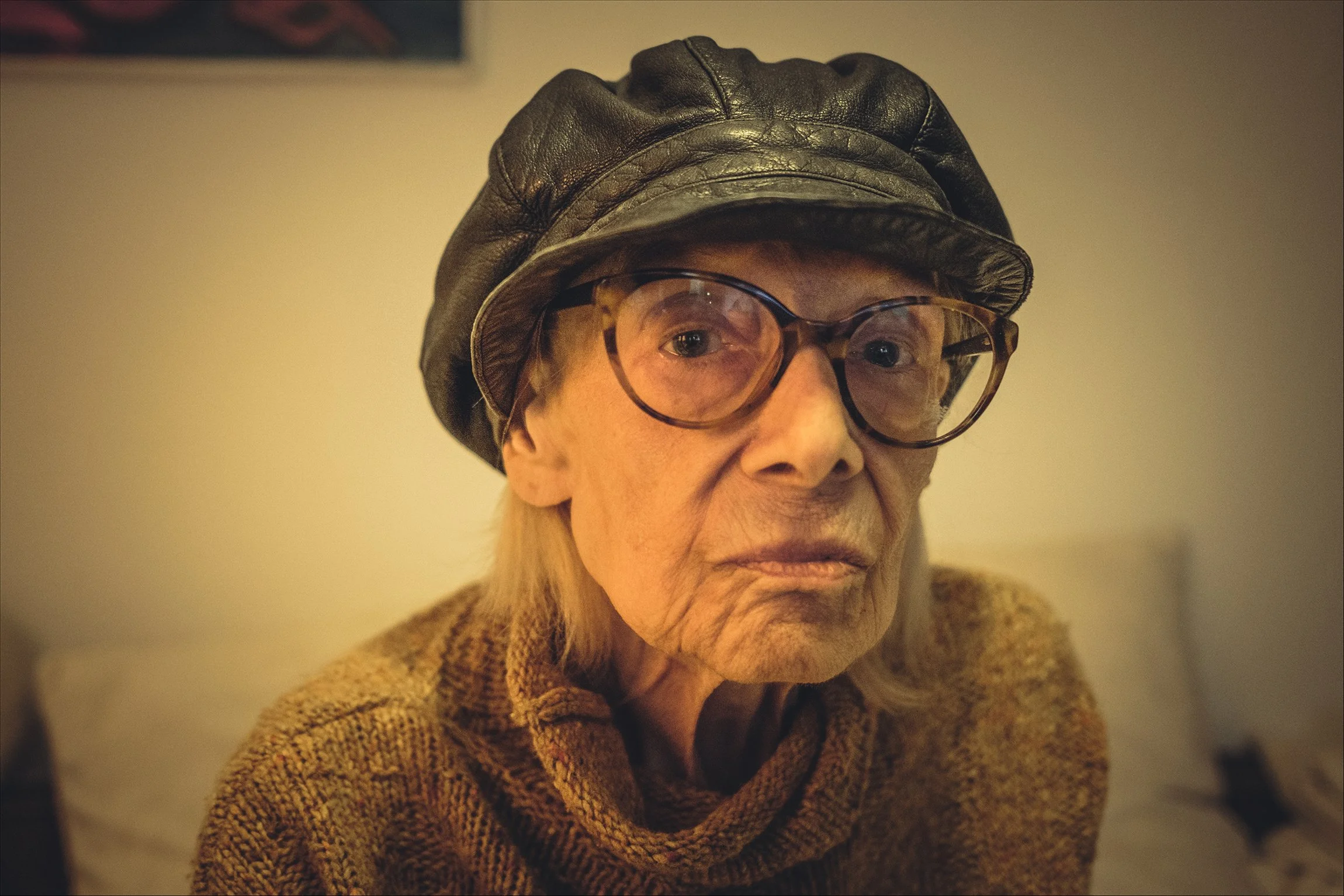 Eine ältere Frau mit Gläsern, schwarzer Ledermütze und kariertem Pullover sitzt in einem Raum mit gelbem Licht.