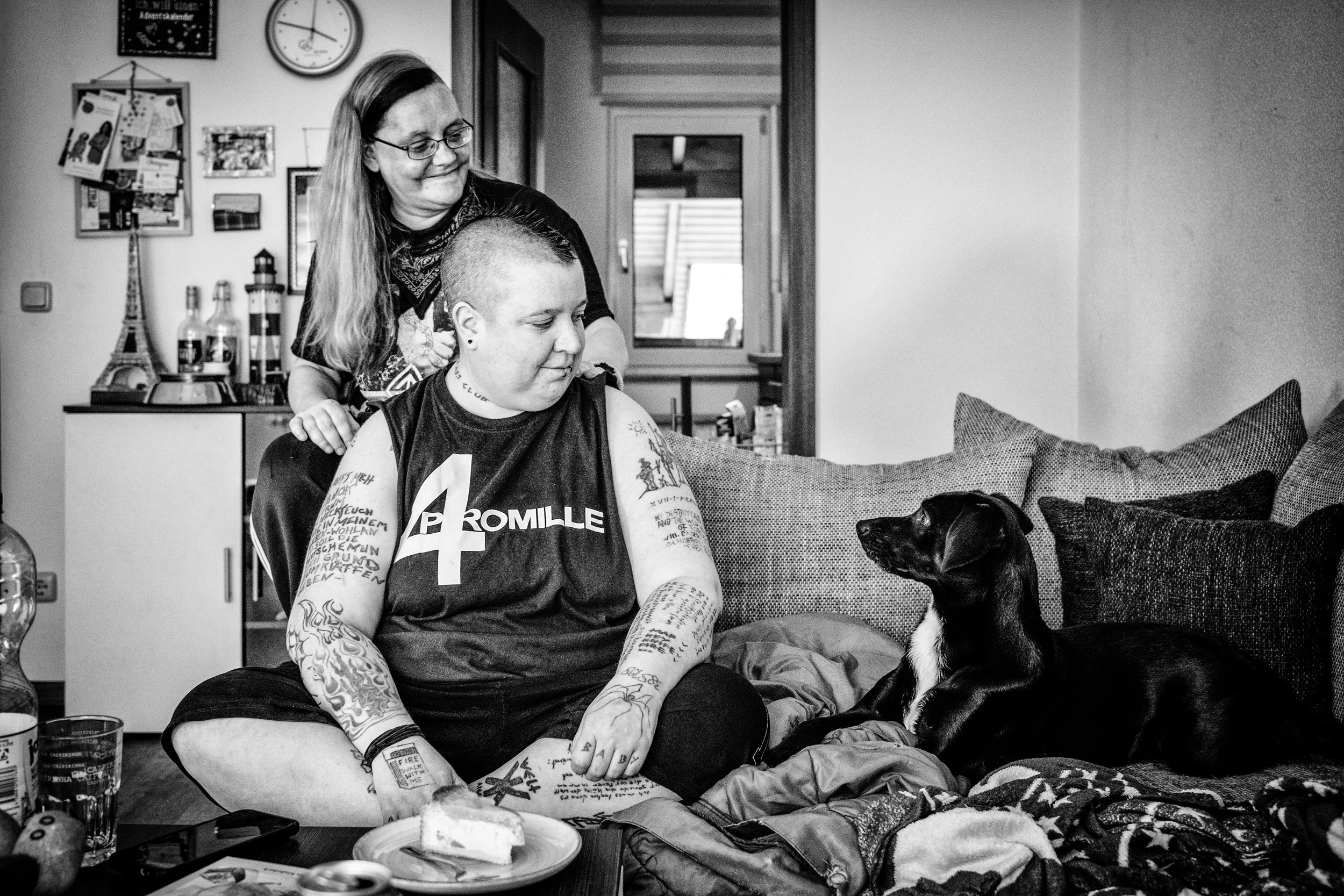 Zwei Frauen, eine sitzend mit Tattoos, und ein Hund sitzen zusammen in einem Wohnzimmer. Eine Frau steht hinter der sitzenden Frau, beide schauen den Hund an.