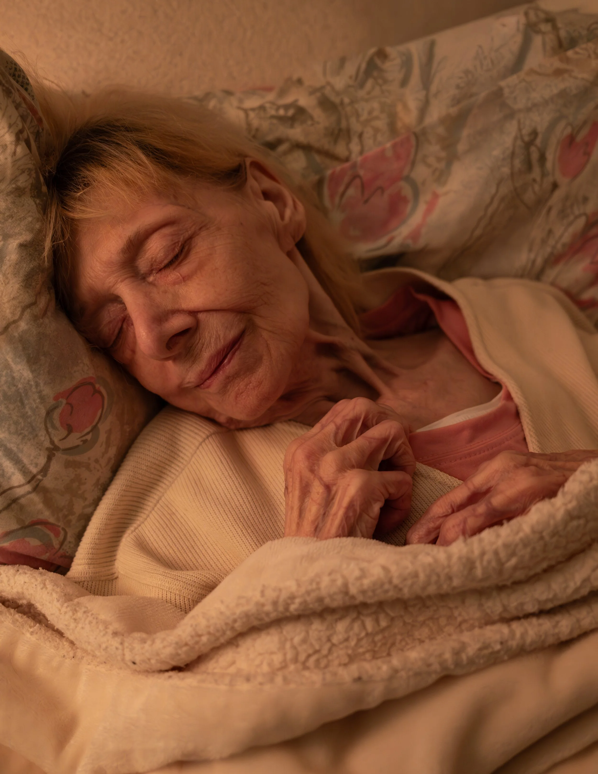 Schläferin, alte Frau, im Bett ruhend, umgeben von Kissen und Decken, warmes Licht.