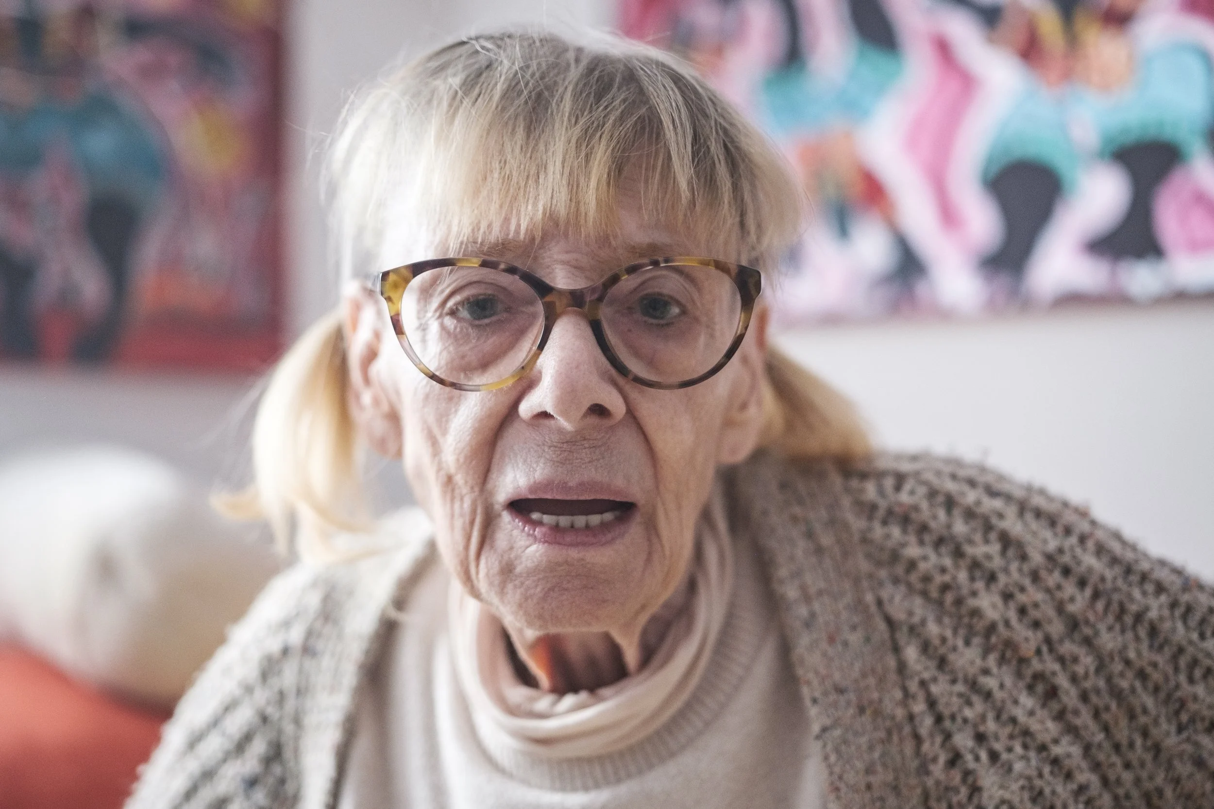Ein älteres Mädchen trägt große, runde Brillen und schaut den Betrachter an, mit einem offenen Mund, im Hintergrund bunte Gemälde.