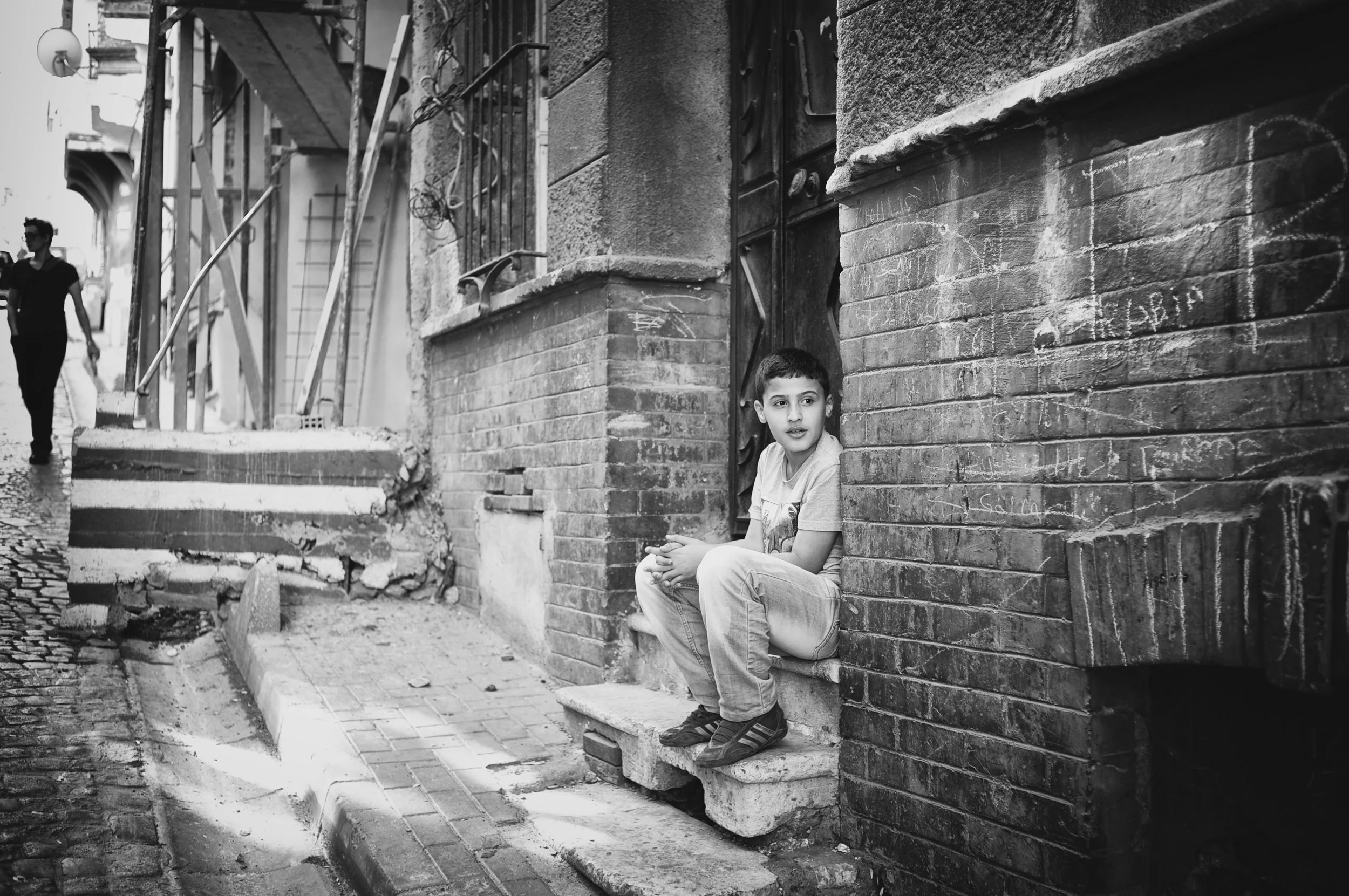 Ein Junge sitzt auf einer kleinen Treppe vor einer alten Ziegelmauer an einem Straßenrand in einer Stadt. Im Hintergrund sind Baugerüste und eine zweite Person zu sehen.