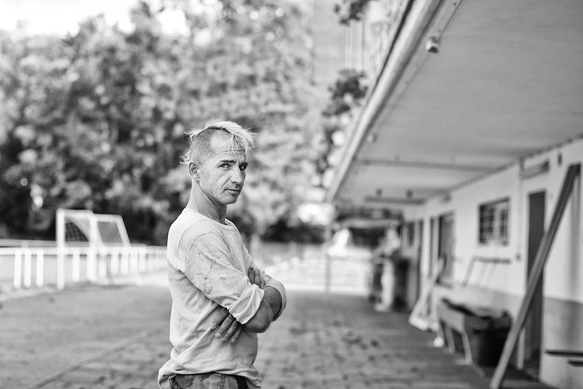 Ein junger Mann mit kurzen, wilden Haaren steht mit verschränkten Armen vor einem Gebäude auf einem Sportplatz, im Hintergrund sind Bäume und Fußballtore sichtbar.