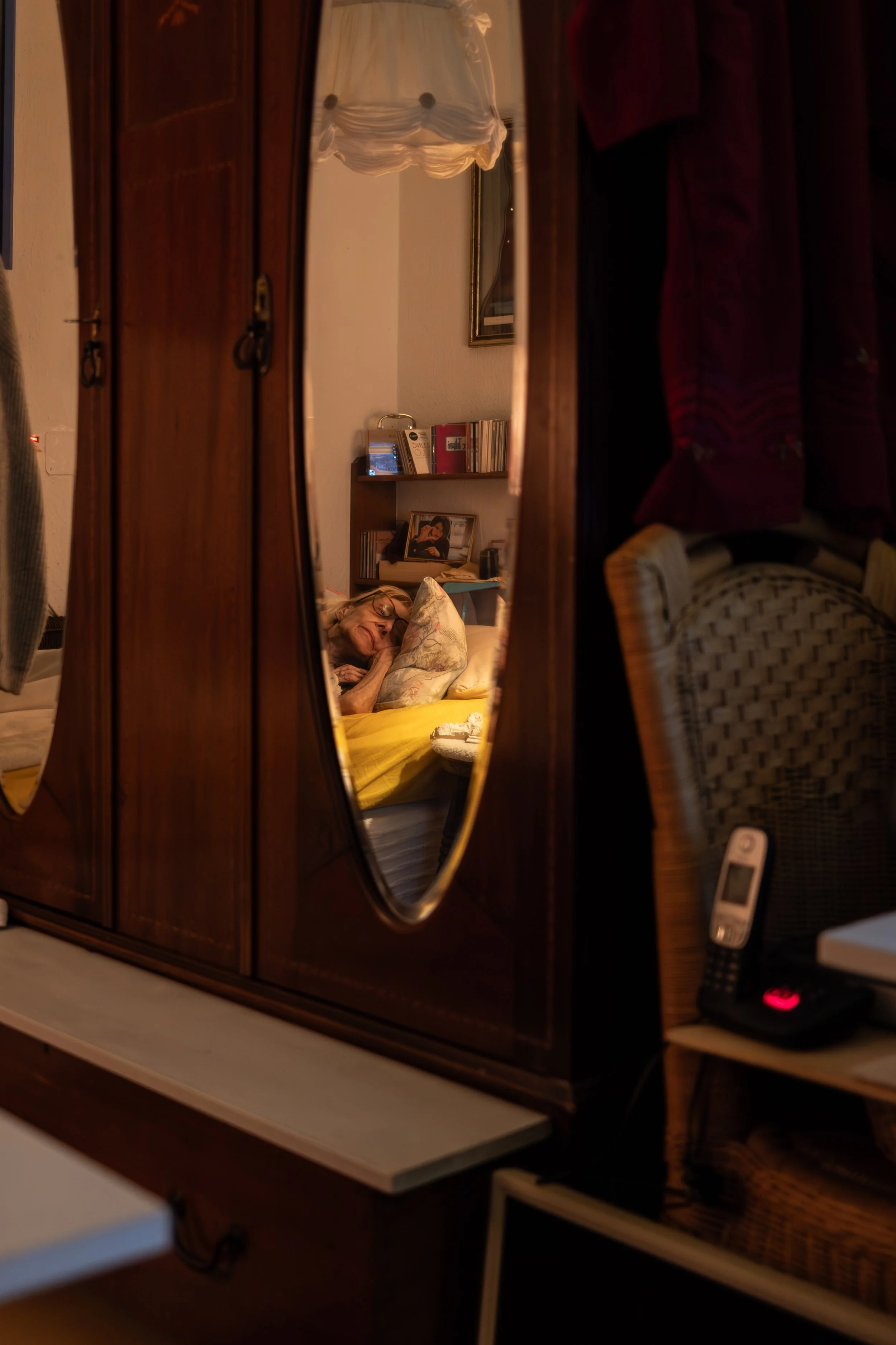 Ein älterer Mann liegt im Bett, gefilmt durch einen Spiegel in einem Schrank. Das Schlafzimmer ist sichtbar, mit Bücherregalen, einem Bild und verschiedenen Gegenständen.