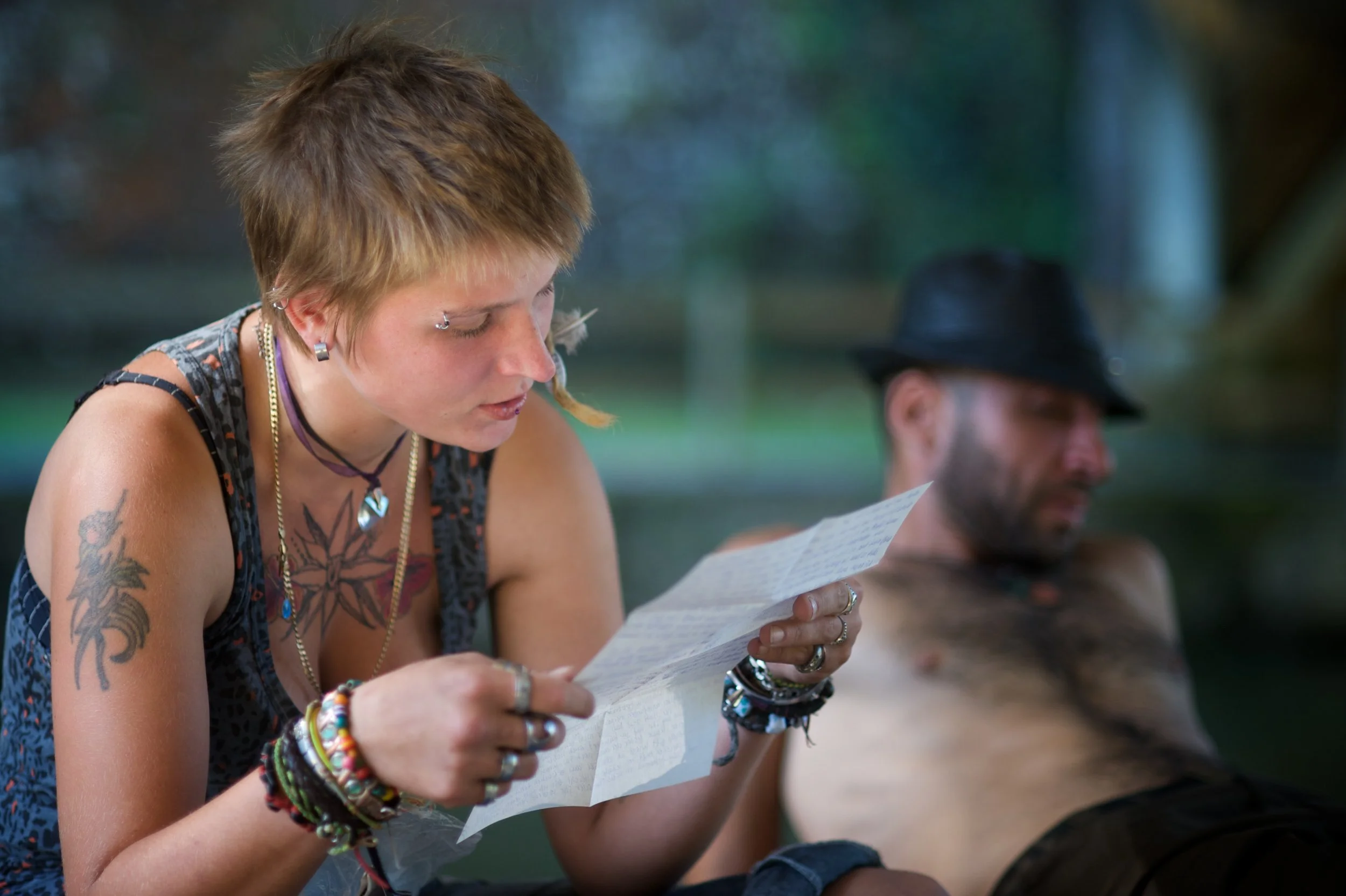Eine junge Frau mit Tattoos liest einen Zettel, während ein Mann im Hintergrund am Wasser entspannt liegt