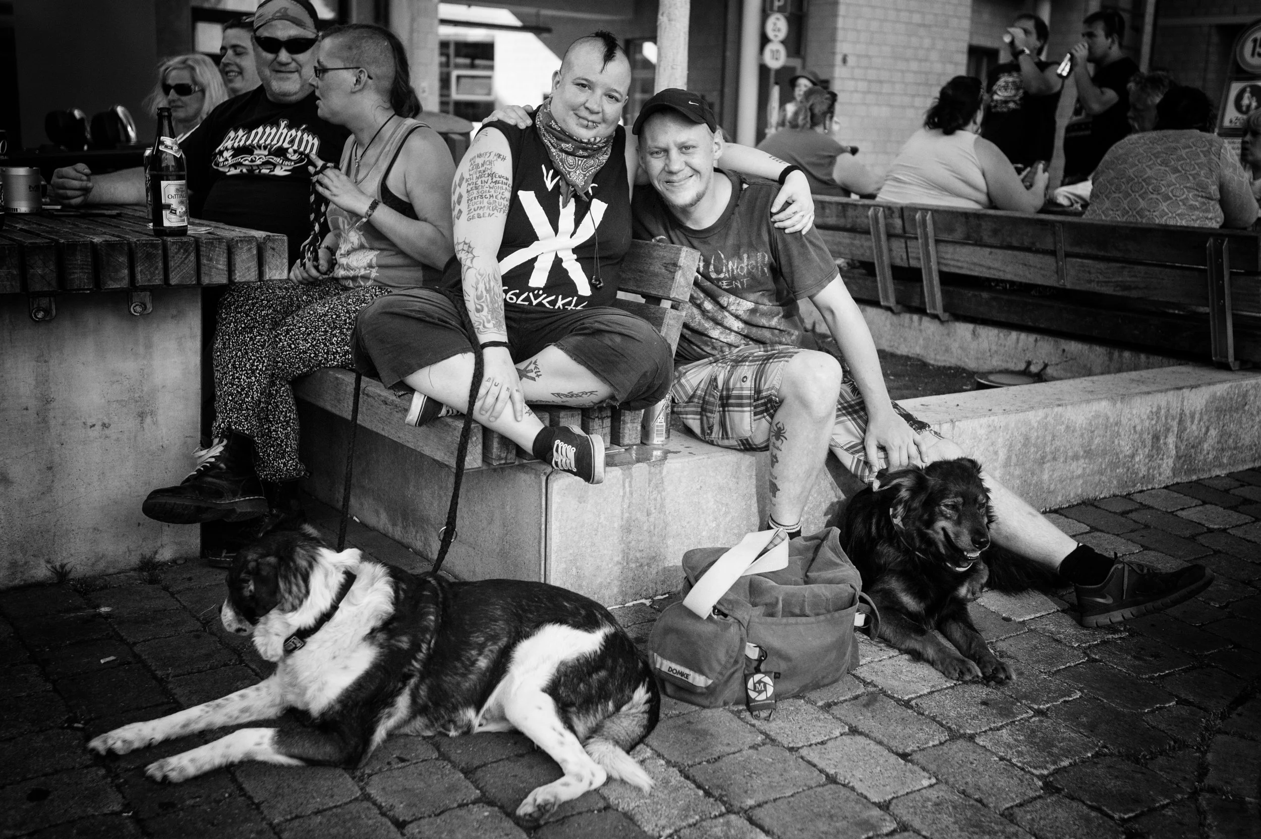 Menschen sitzen an einem öffentlichen Platz, einige im Vordergrund, darunter eine Gruppe von zwei jungen Menschen mit Tattoos auf der rechten Seite, die auf einer Bank sitzen, neben ihnen liegen zwei Hunde. Im Hintergrund sind weitere Menschen an Tis