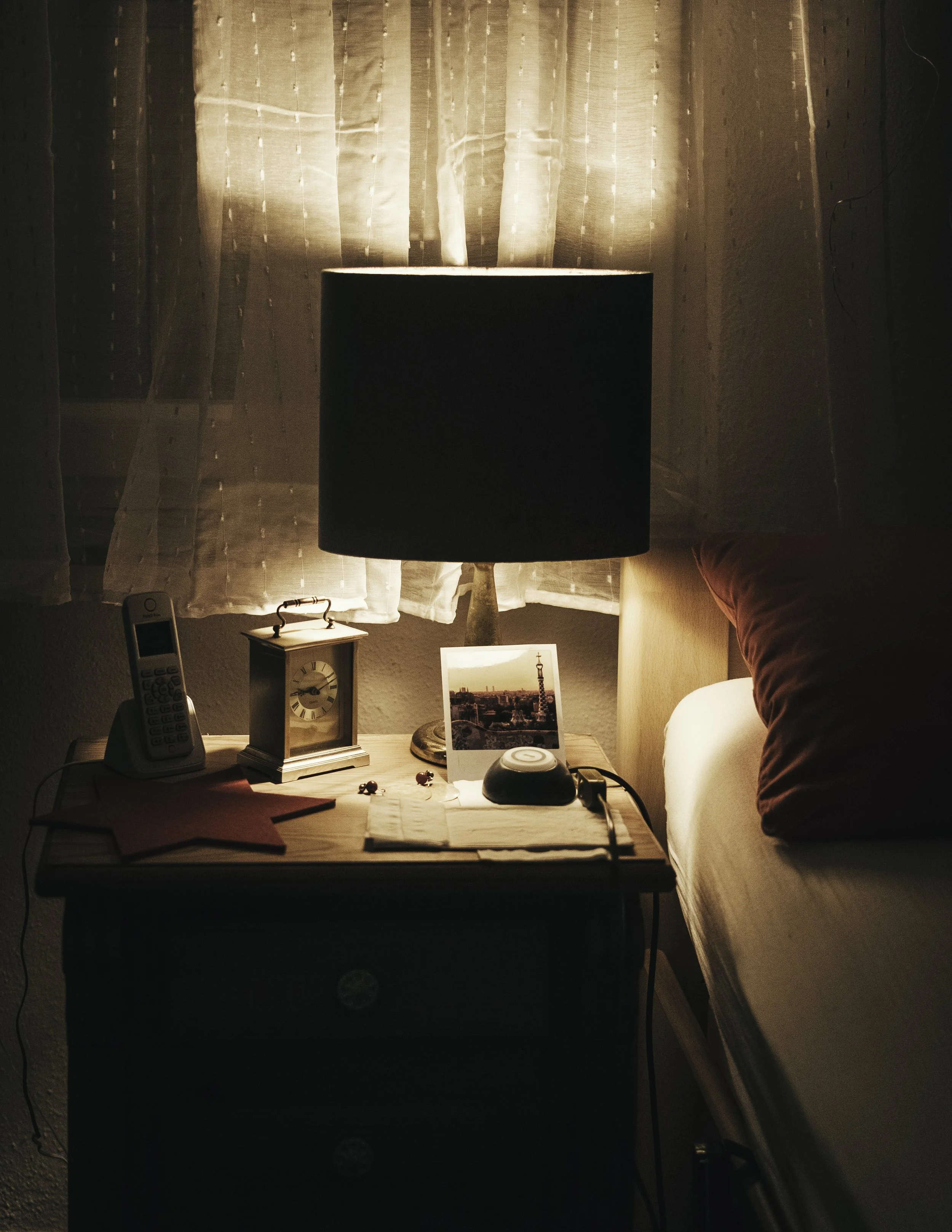 Nachts beleuchteter Nachttisch neben einem Bett mit rotem Kissen, auf dem Nachttisch stehen eine kleine Uhr, ein Telefon, eine mit einem Foto versehenes Karten, eine frühe Schnecke, die auf dem Nachttisch liegt, und eine Lampe, die den Raum beleuchte