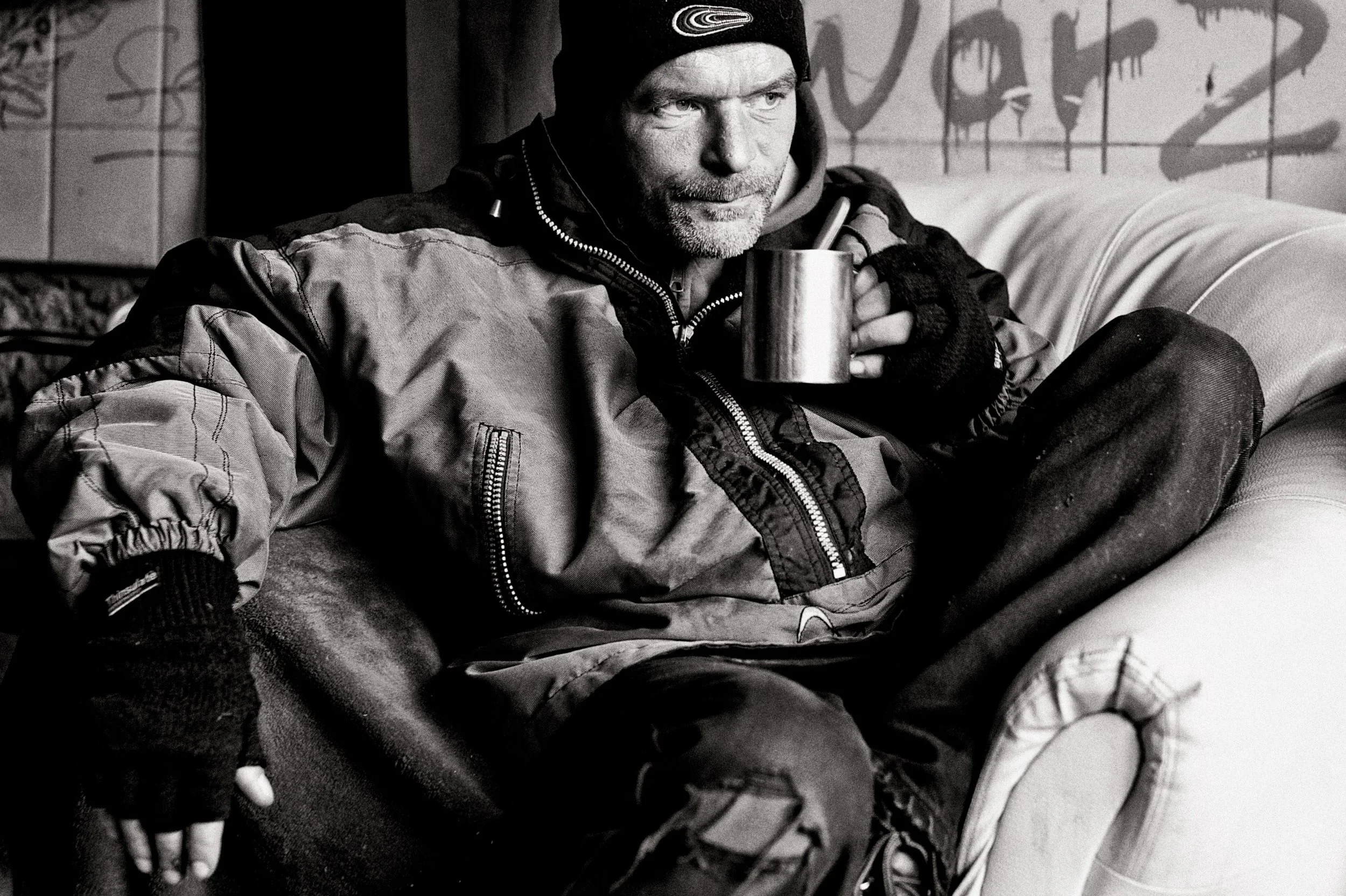 Ein Mann sitzt auf einer Couch, trägt eine Jacke, Mütze und Handschuhe, und hält eine Tasse.