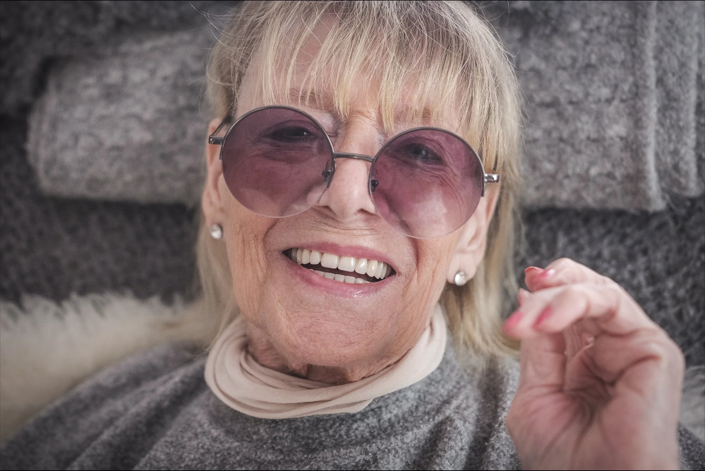 Lächelnde ältere Frau mit großen, rosa Sonnenbrillen, grauem Pullover und weißem Schal, sitzend vor einer Steinmauer.